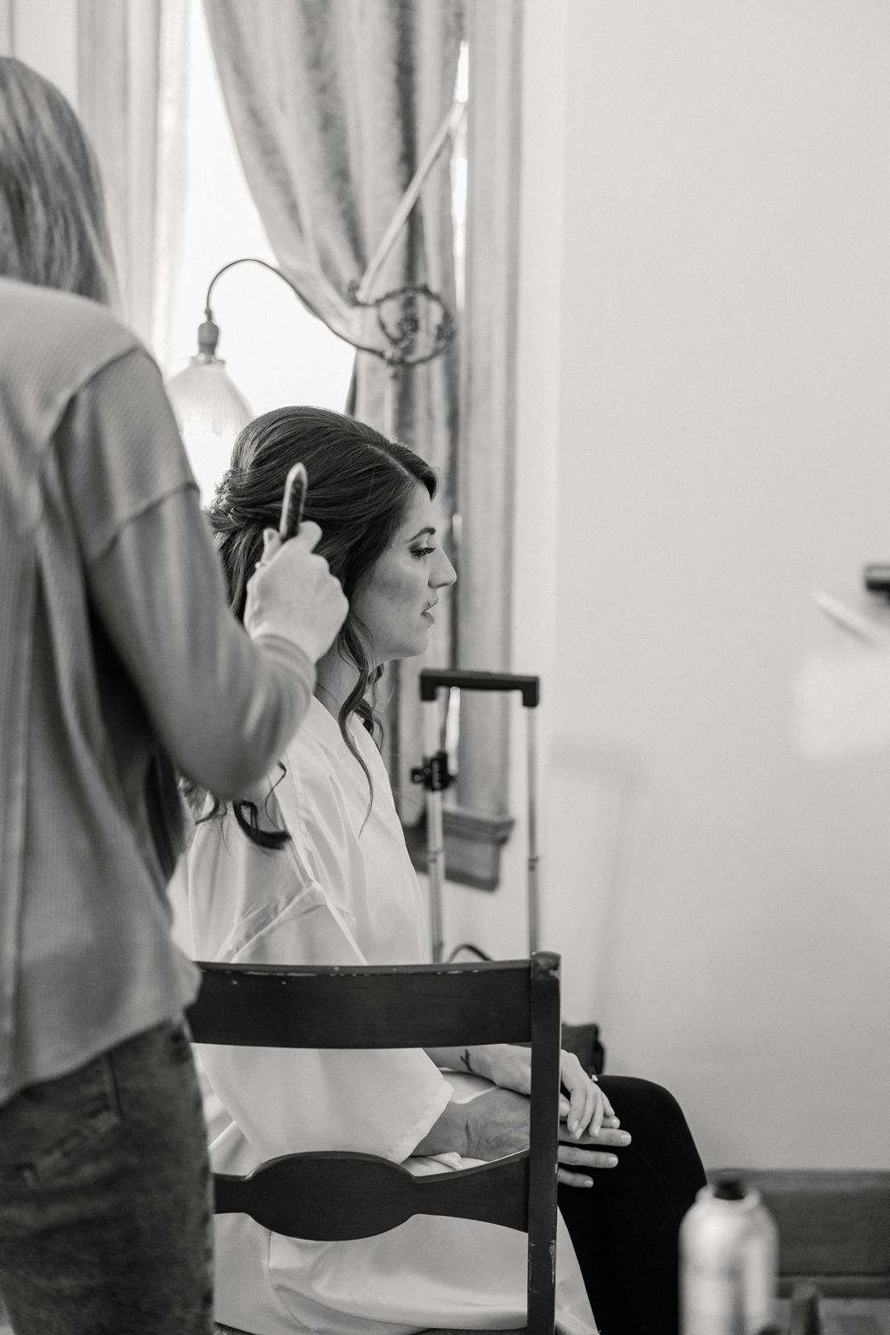 Black and white photo showing wedding day hair styling preparation in front of a window.