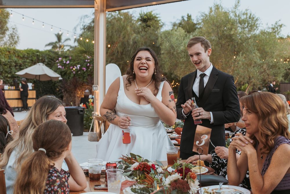 Joyful celebration at an outdoor wedding reception with guests laughing around a decorated dining table at River Garden Weddings and Events.