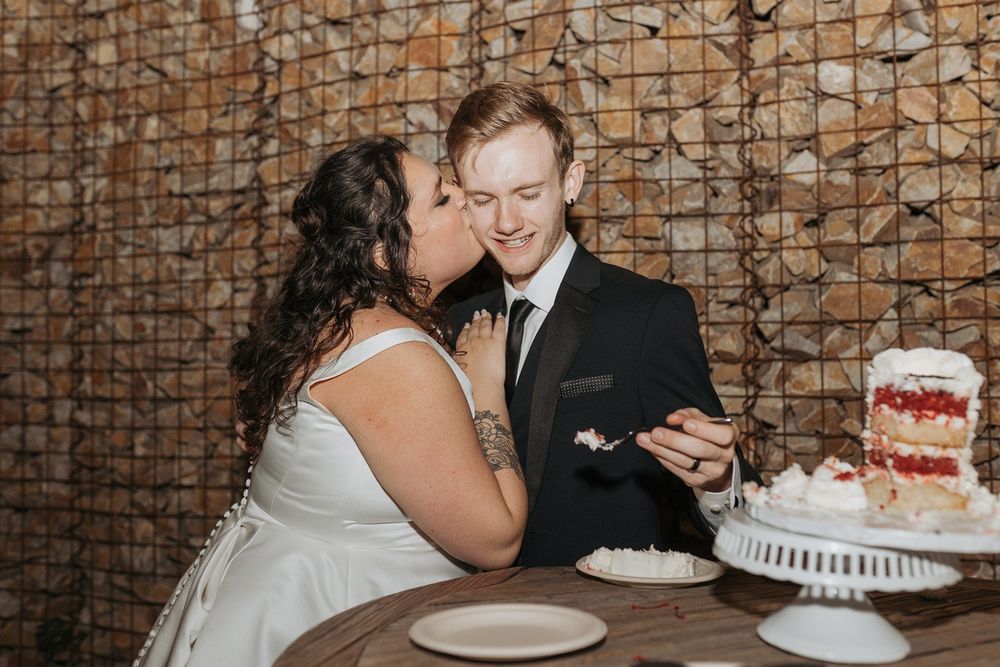 A couple sharing sweet moments and eating wedding cake at their reception with a stone wall backdrop at River Garden Weddings and Events.