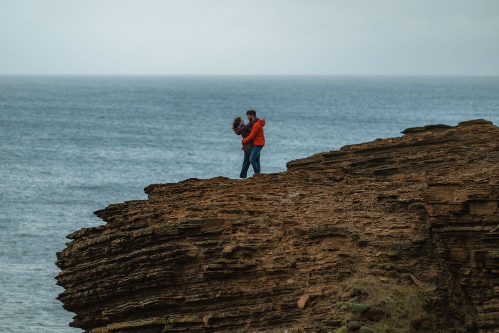 Two people stand on rugged coastal cliffs overlooking the ocean waves crashing against rocky shores.