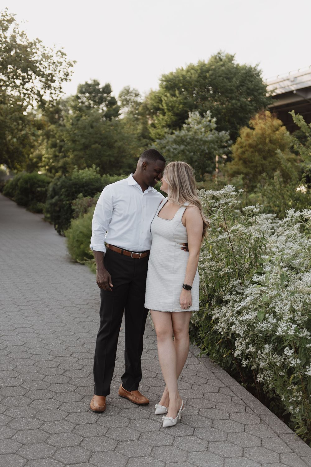 A couple shares a romantic moment on a stone path surrounded by blooming white wildflowers at sunset.