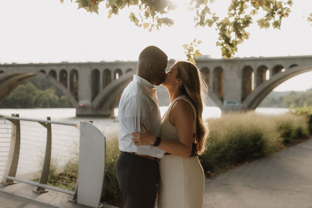 A couple embraces during sunset with trees and architecture in the background.