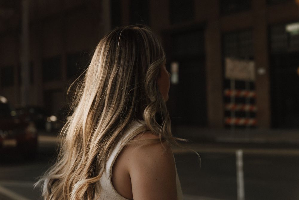 A person with long blonde hair stands in a dark room with moody lighting and backlit atmosphere.