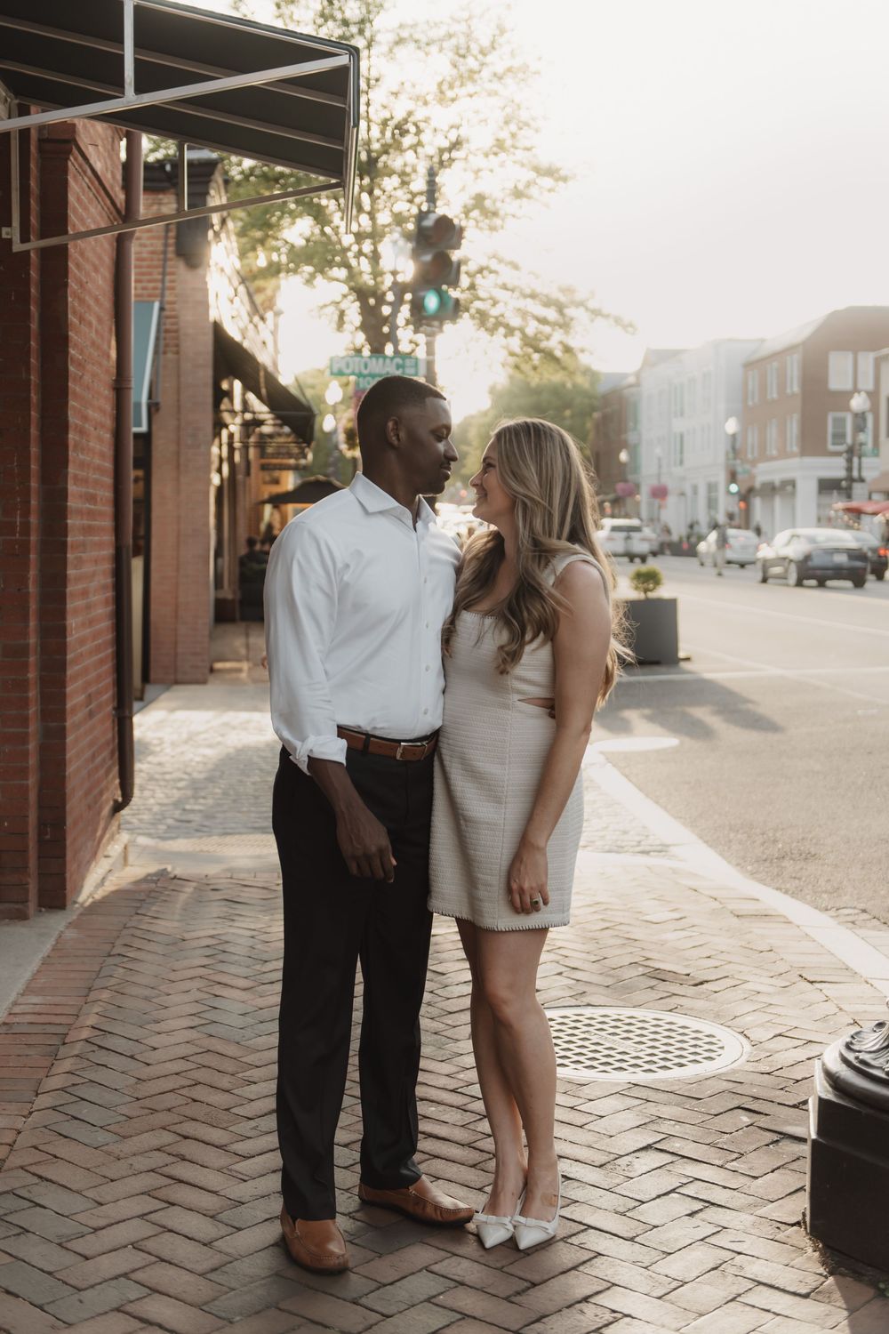 Couple stands together on brick sidewalk during golden hour in urban setting.
