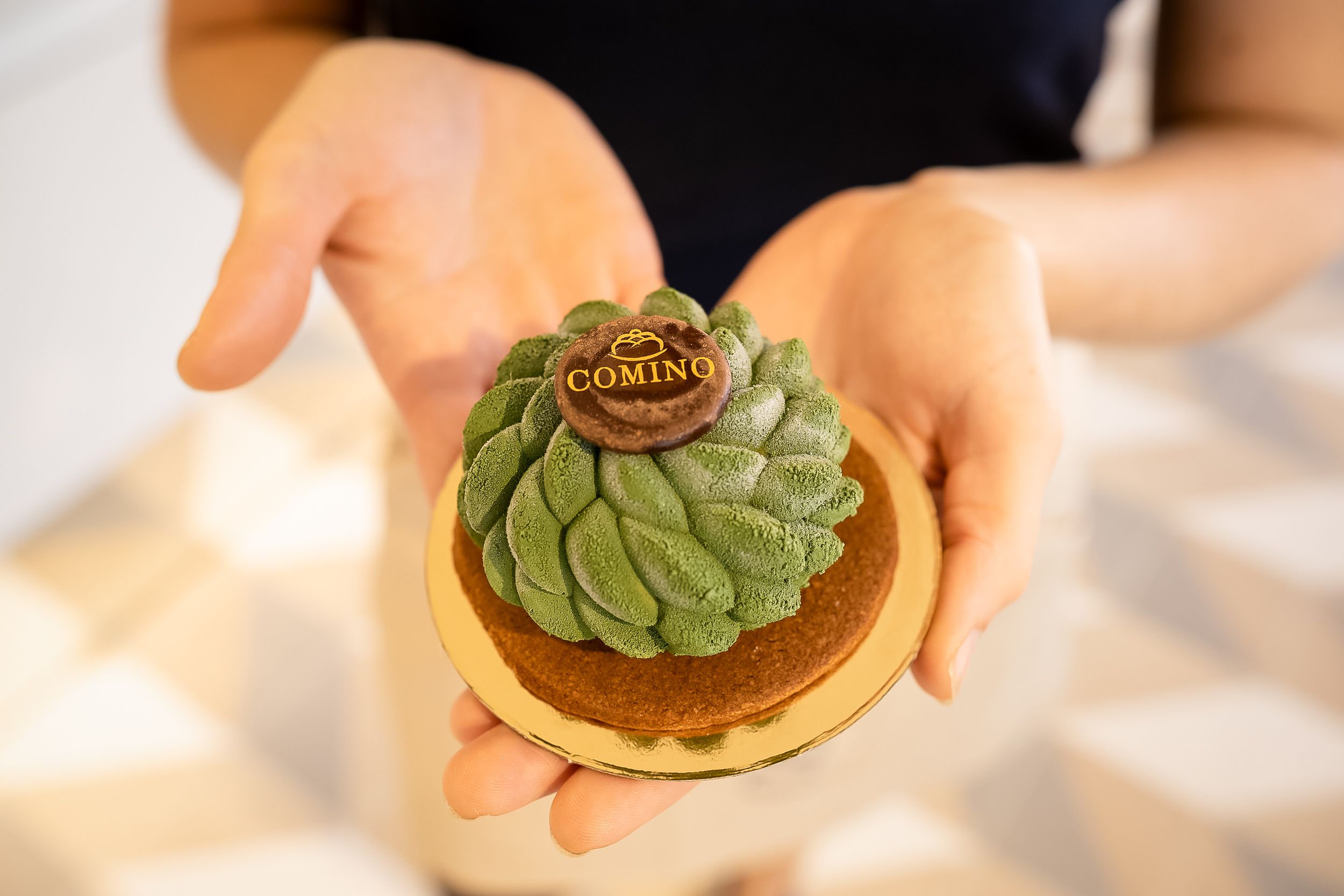 Hands holding a beautifully crafted matcha green tea tart with intricate leaf design and gold accent on top.