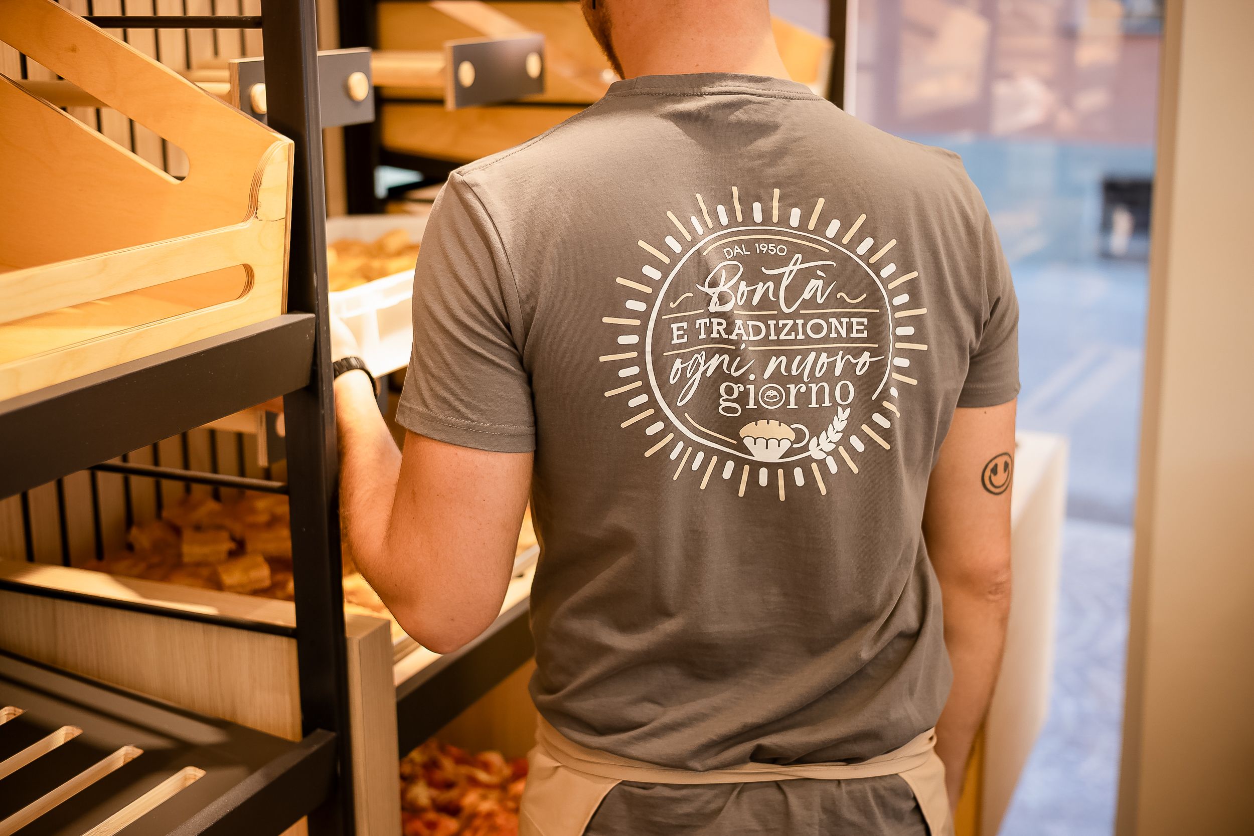 Person wearing a gray t-shirt with text logo stands in front of wooden shelves in a retail setting.