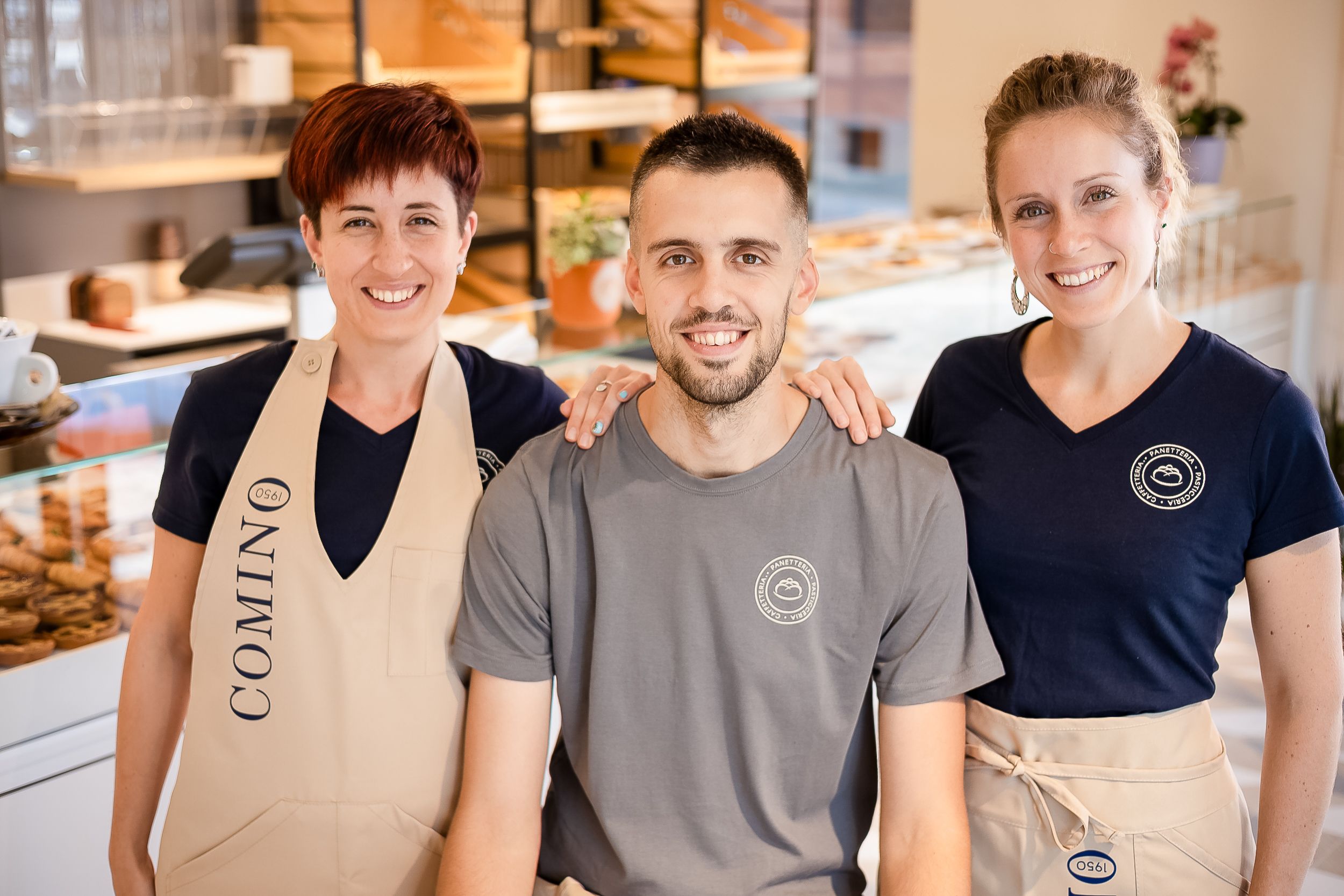 Smiling employees in beige aprons and casual attire stand together in a bright modern cafe setting.