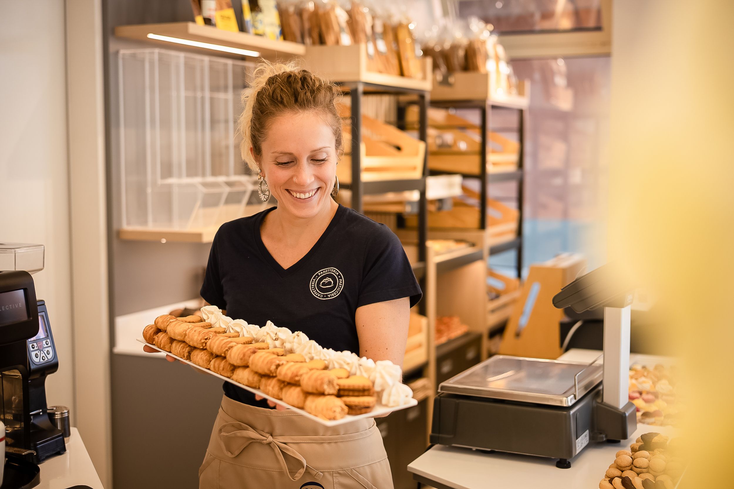 A bakery worker arranges fresh baked goods on display shelves in a warm, inviting retail setting.