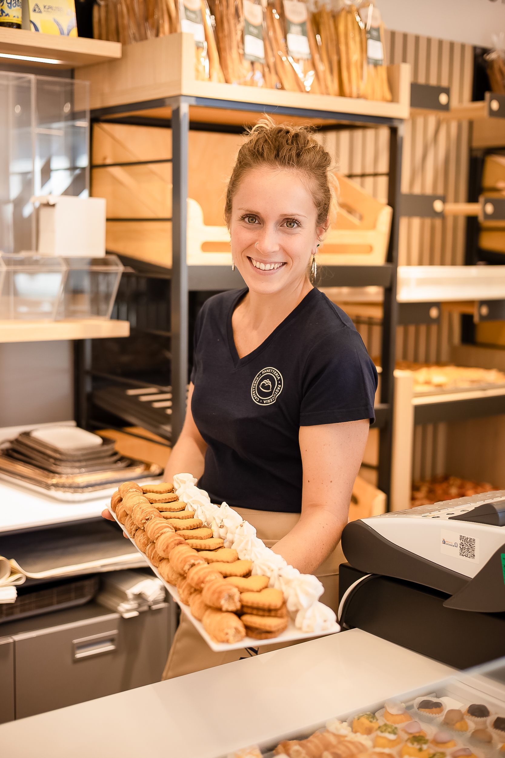 A bakery employee in a black uniform shirt displays a tray of fresh baked goods at a retail counter.