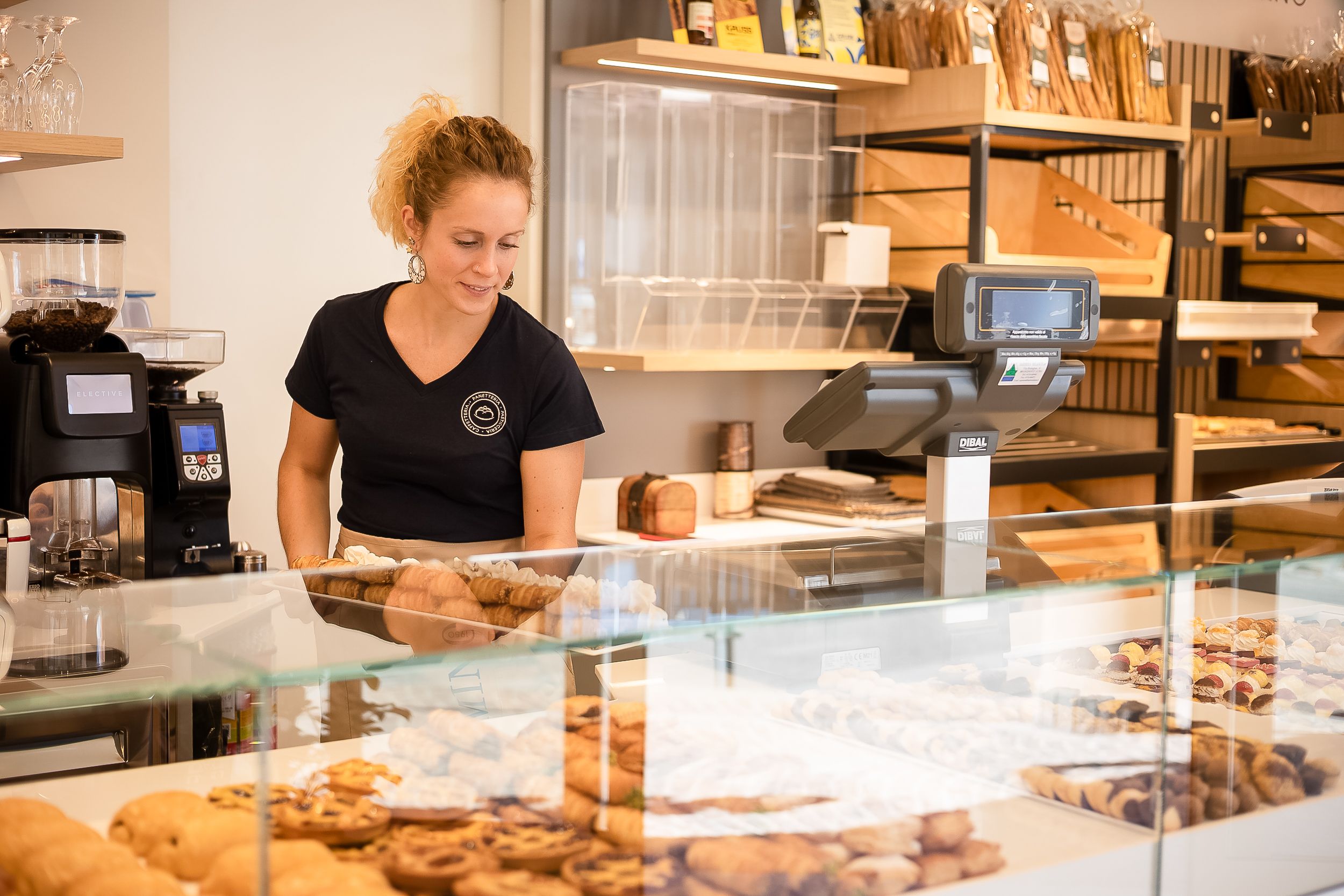 A bakery employee arranges pastries in a glass display case while working behind the counter.