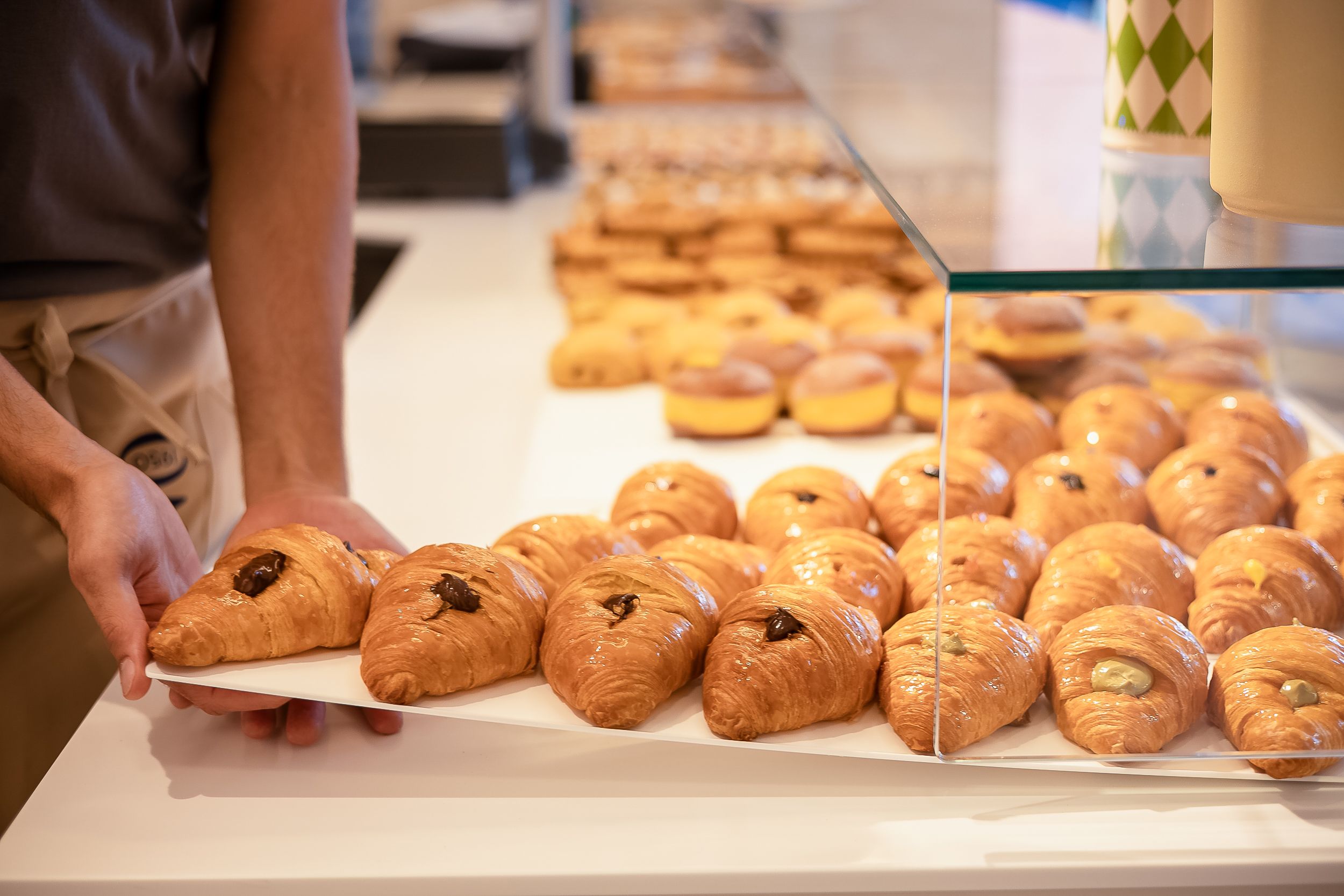 Fresh bagels lined up on a bakery counter with various toppings and textures visible in warm lighting.
