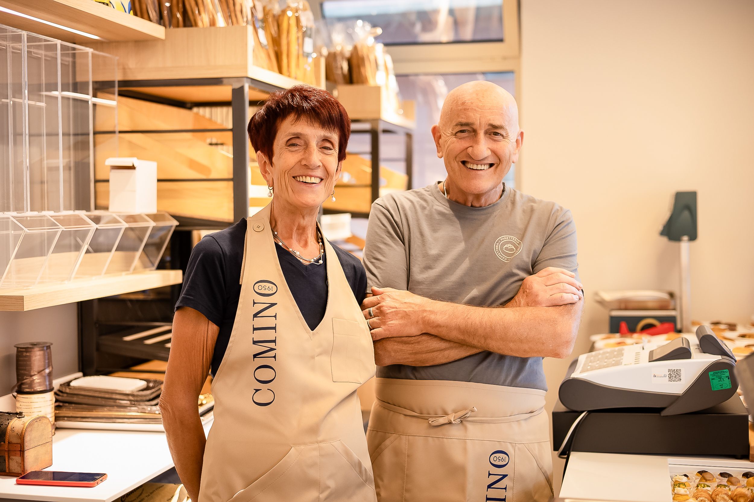Two bakers wearing Oonini aprons stand together smiling in a commercial kitchen workspace.