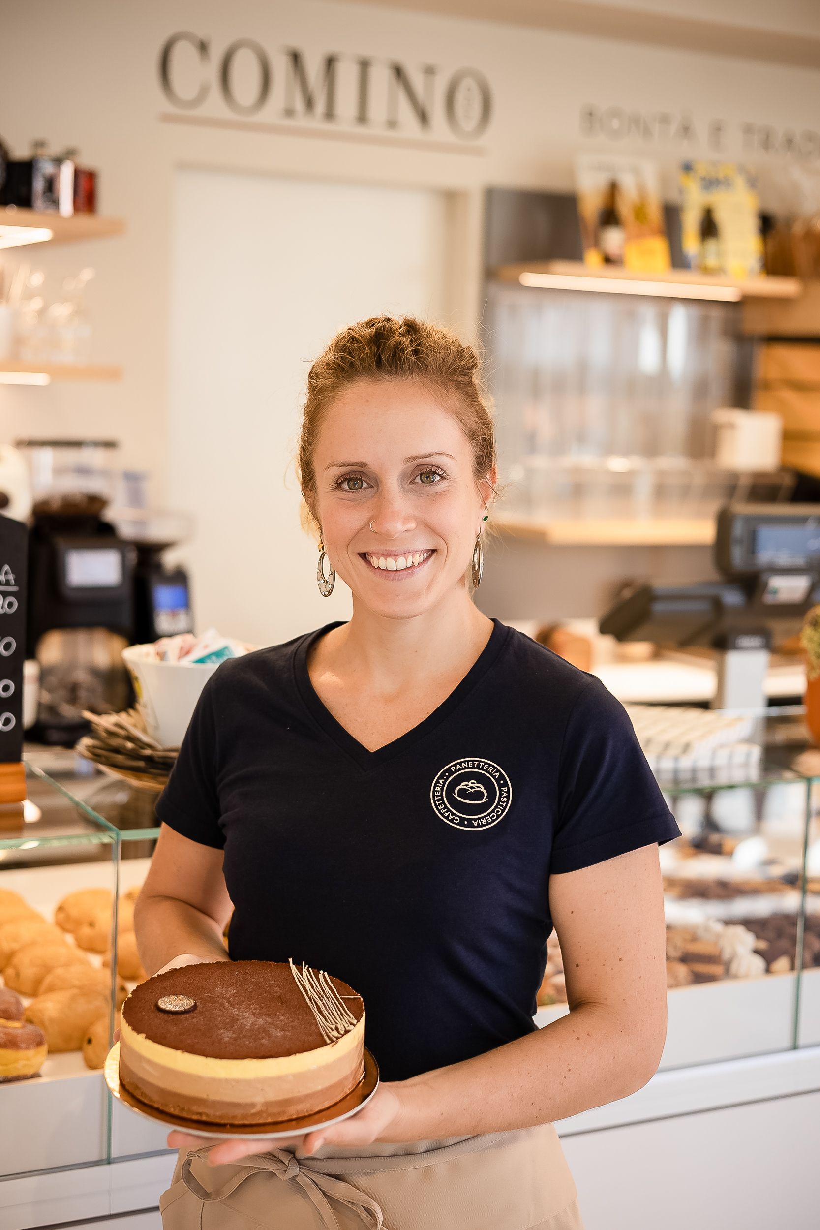Staff member in navy uniform smiles while working behind counter at Comino bakery and restaurant.