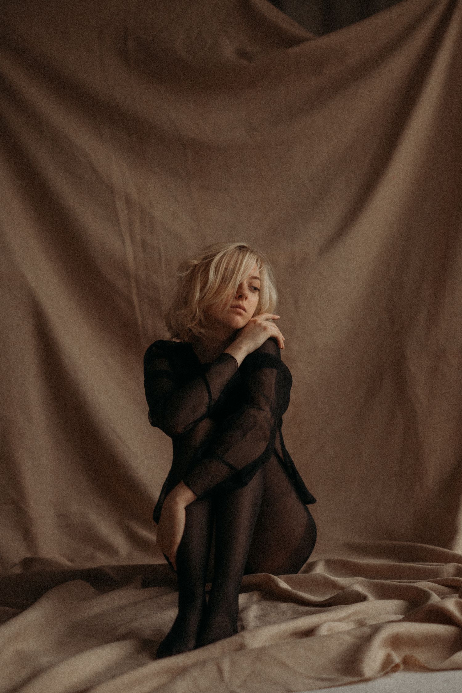 A woman in a black outfit poses artistically against a beige backdrop in a moody boudoir photoshoot.