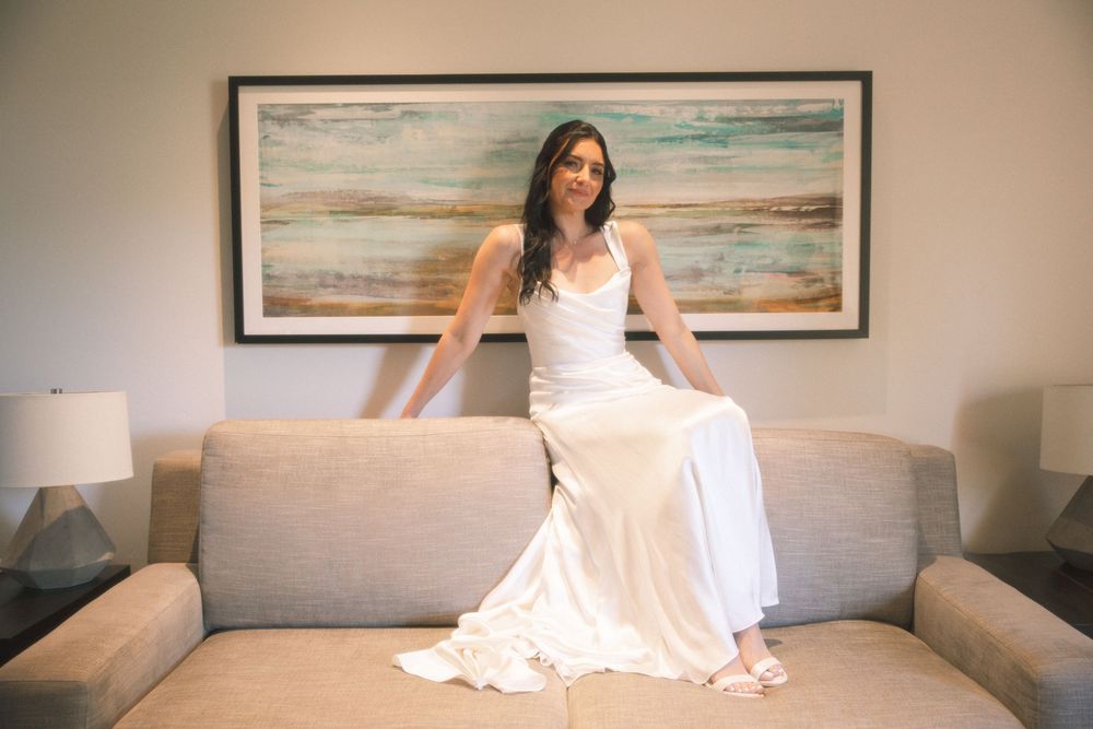 A person in a flowing white dress sits elegantly on a beige couch with abstract artwork displayed on the wall behind.
