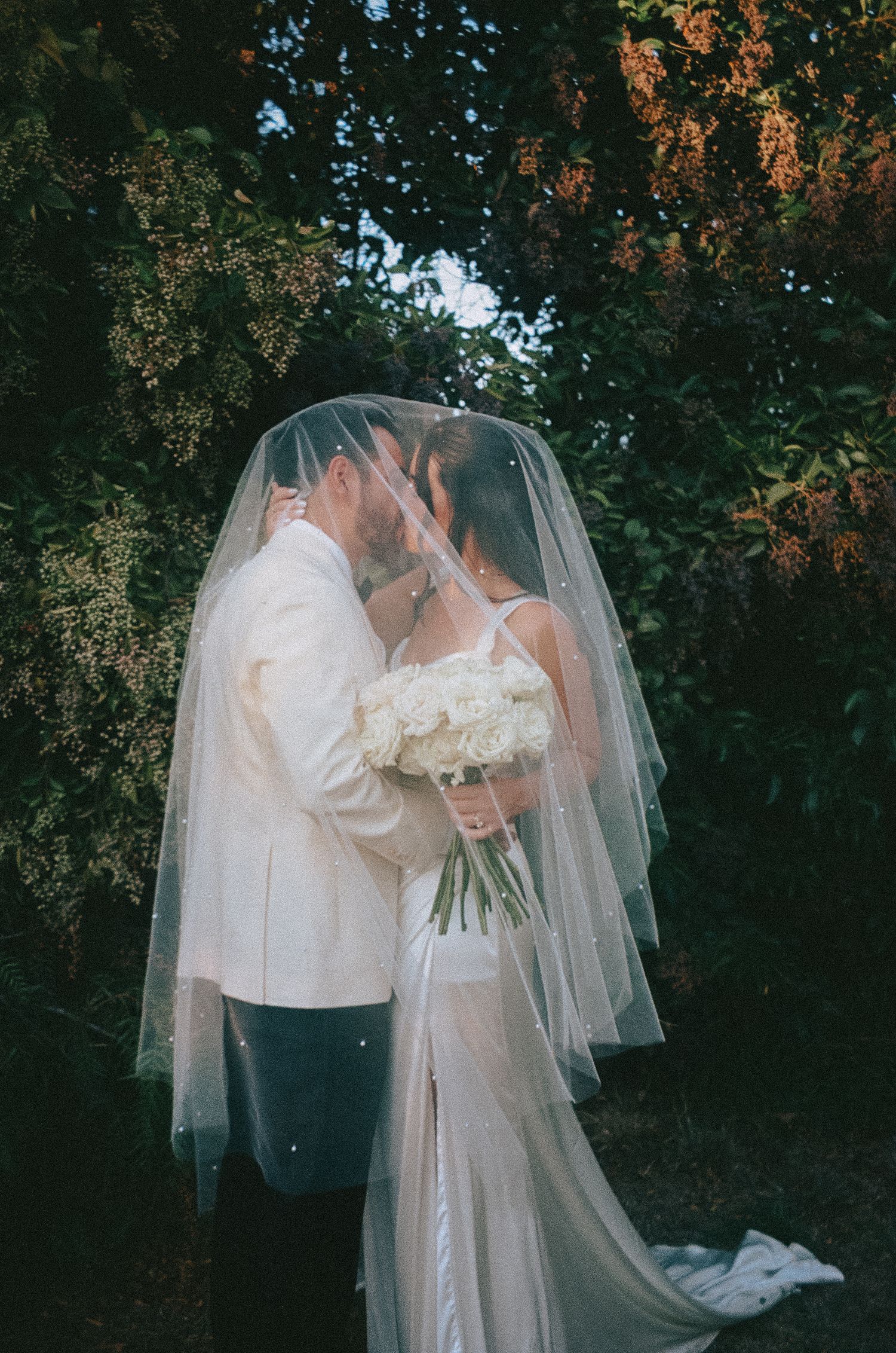 A romantic wedding couple shares intimate moments in a garden setting with autumn foliage in the background.