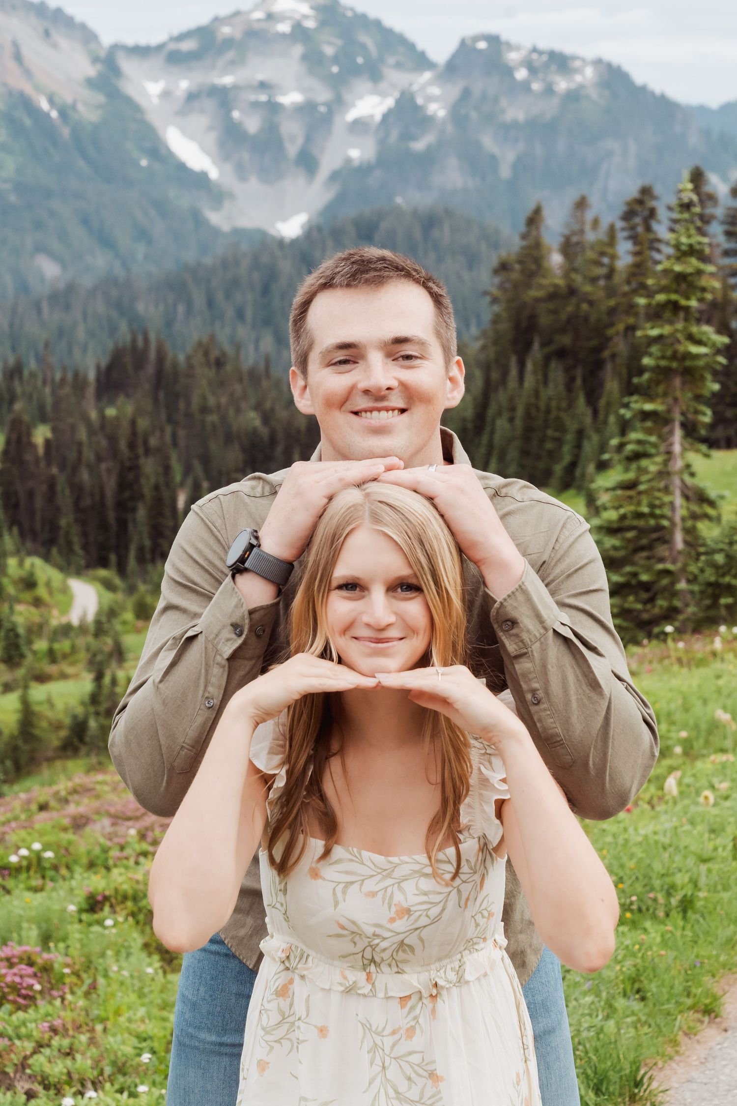 A couple poses playfully in a scenic mountain meadow with snow-capped peaks and evergreen forest in the background.