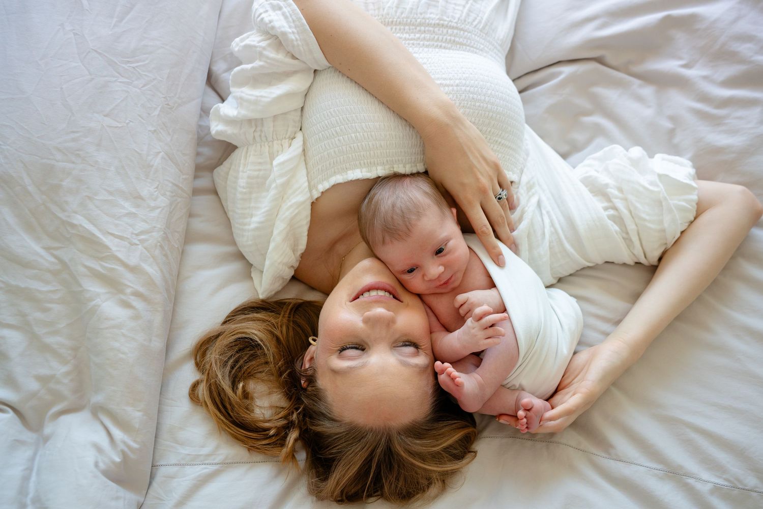 A tender moment captured from above as a mother and baby lie together on white bedding.