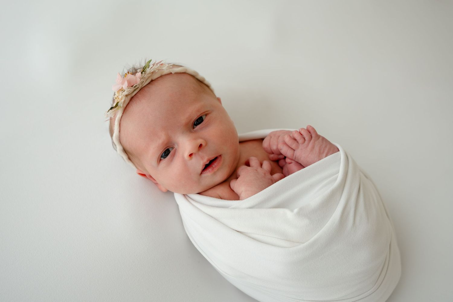 Newborn baby swaddled in white fabric against light background.