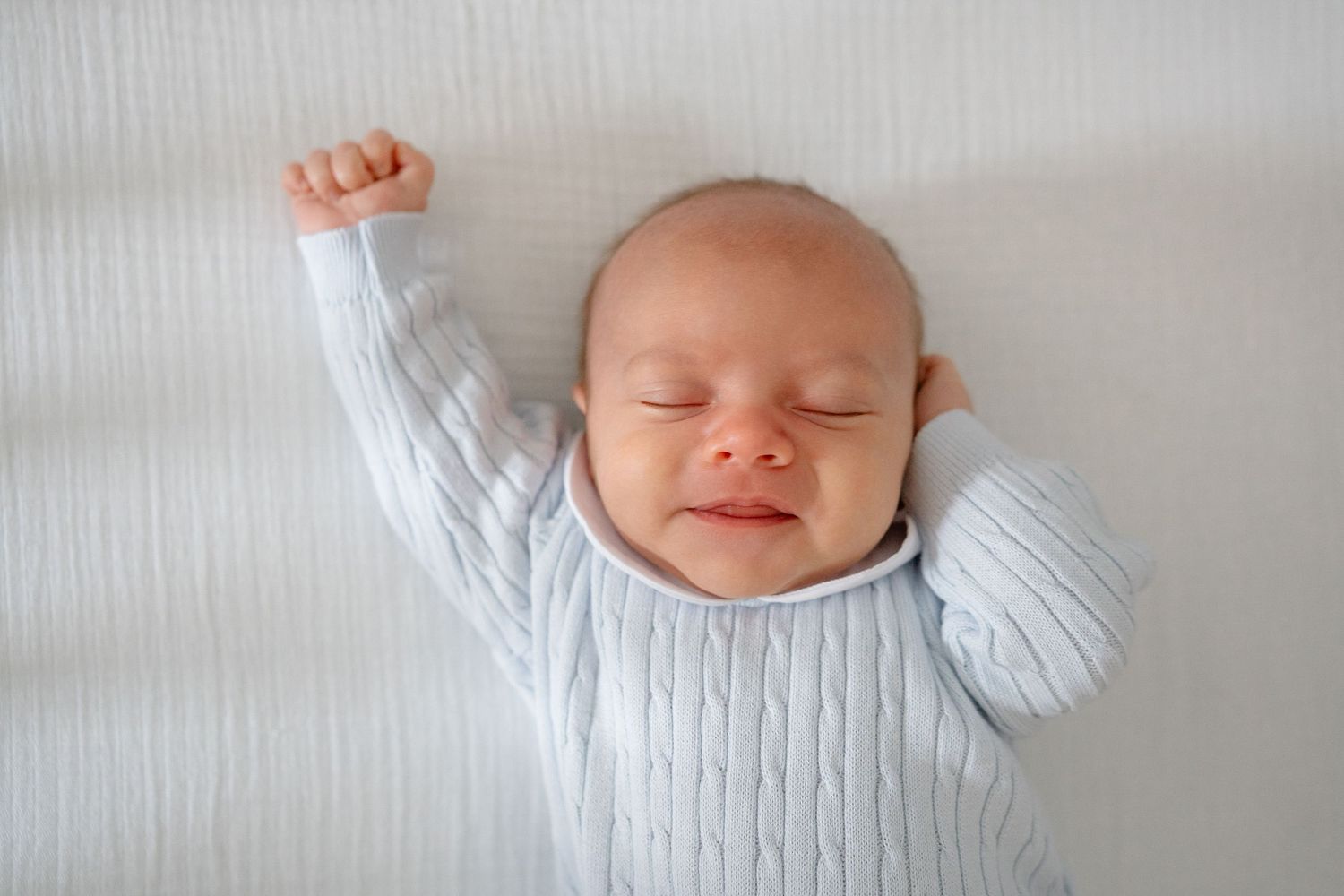 A sleeping newborn baby wearing a white onesie lies peacefully on white bedding.