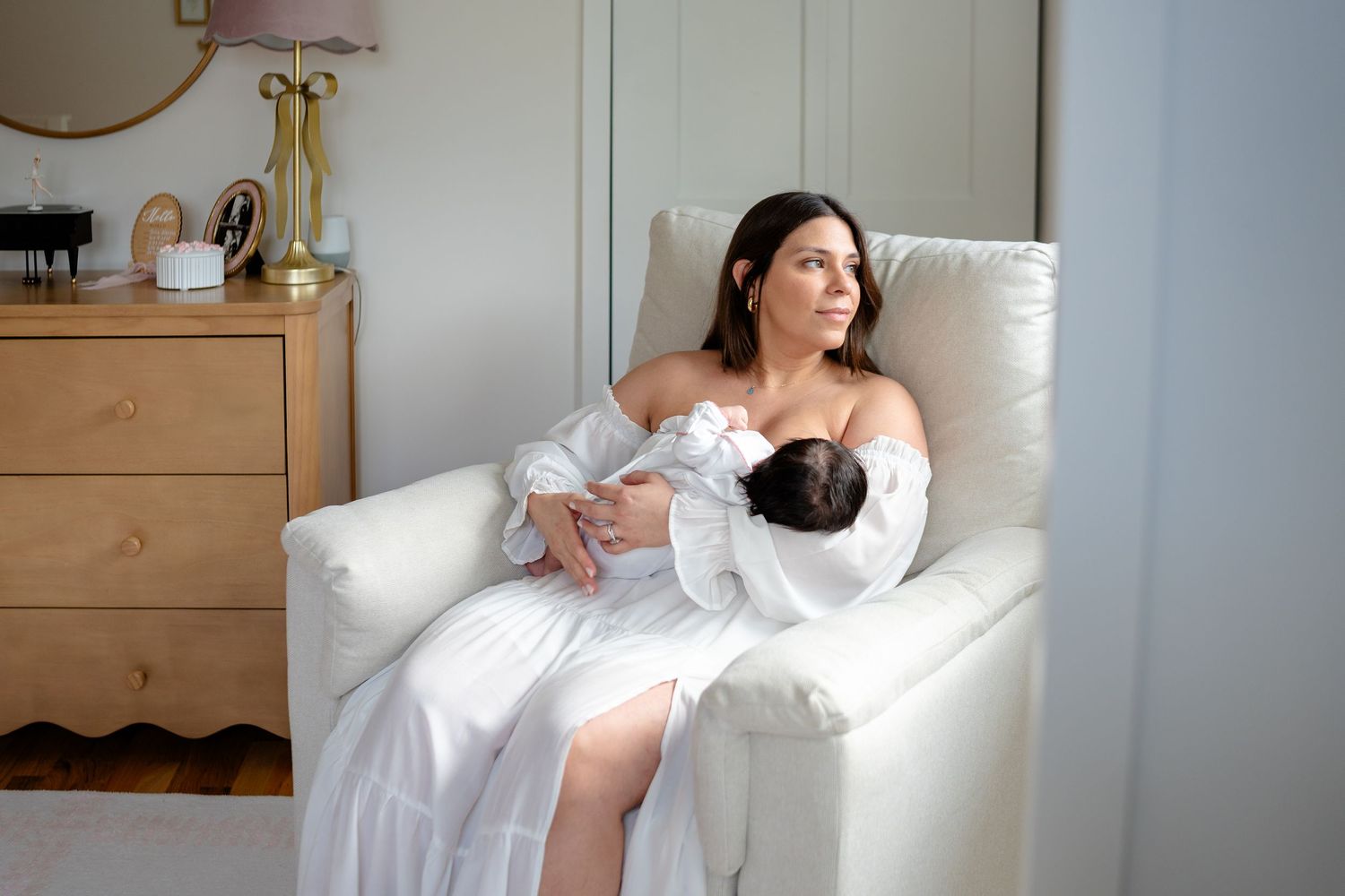 A person in a white dress sits in a cozy armchair holding a sleeping baby in a tranquil bedroom setting.