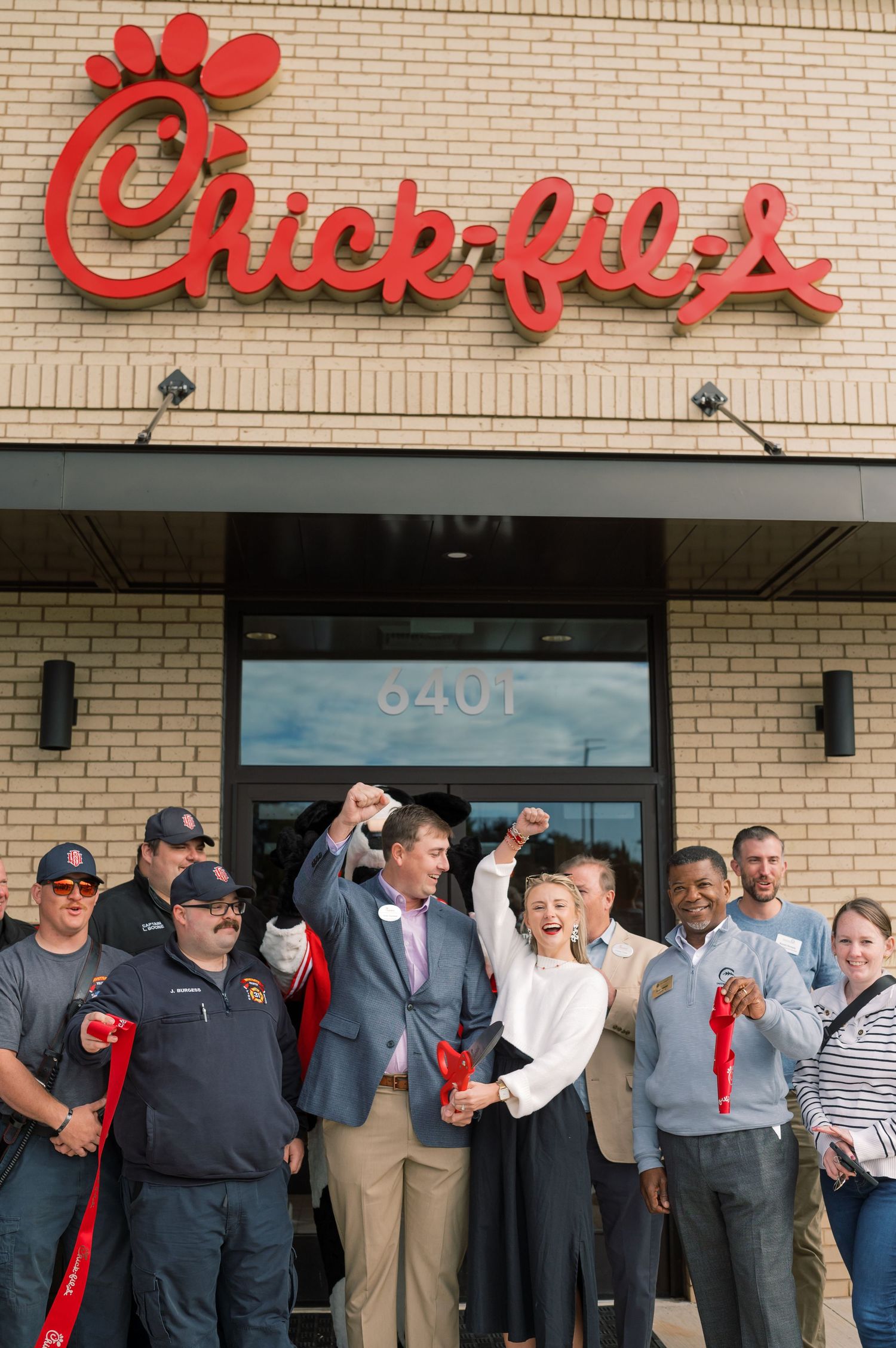 Group celebrating grand opening outside Chick-fil-A restaurant with red ribbon cutting ceremony.