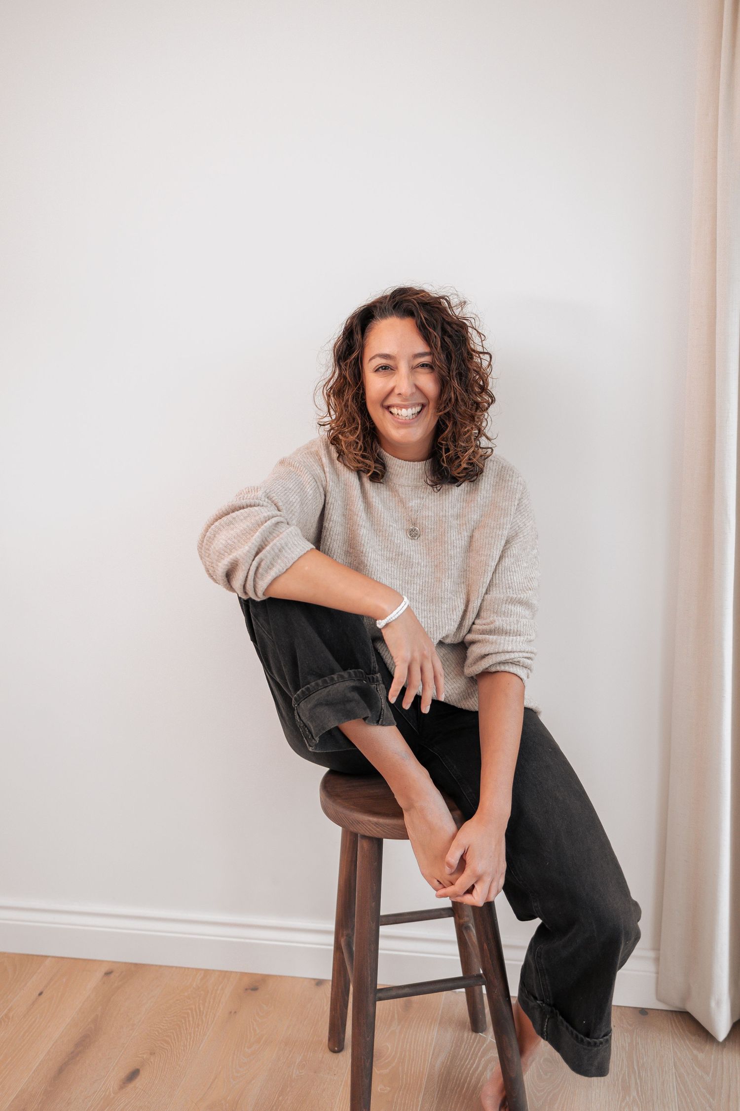 Woman in casual grey sweater posing playfully on wooden chair against white wall. Photography in Buckinghamshire.