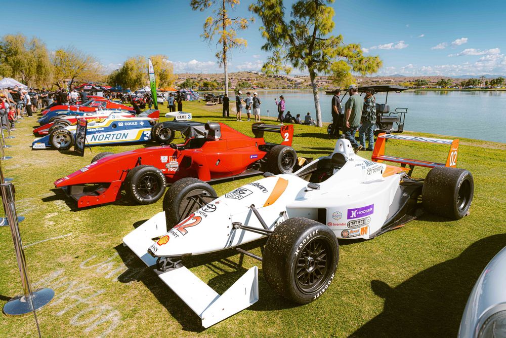 Vintage race cars and Formula One vehicles displayed on grass at an outdoor automotive exhibition near a lake.