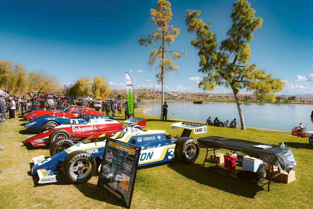 Classic cars line up along a grassy lakeside area under blue skies during a vintage automobile show.