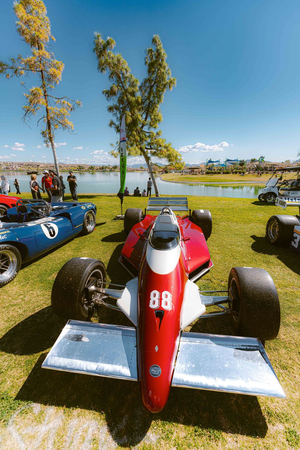 Classic race cars displayed at an outdoor vintage car show on a sunny day by a lake with trees in the background.
