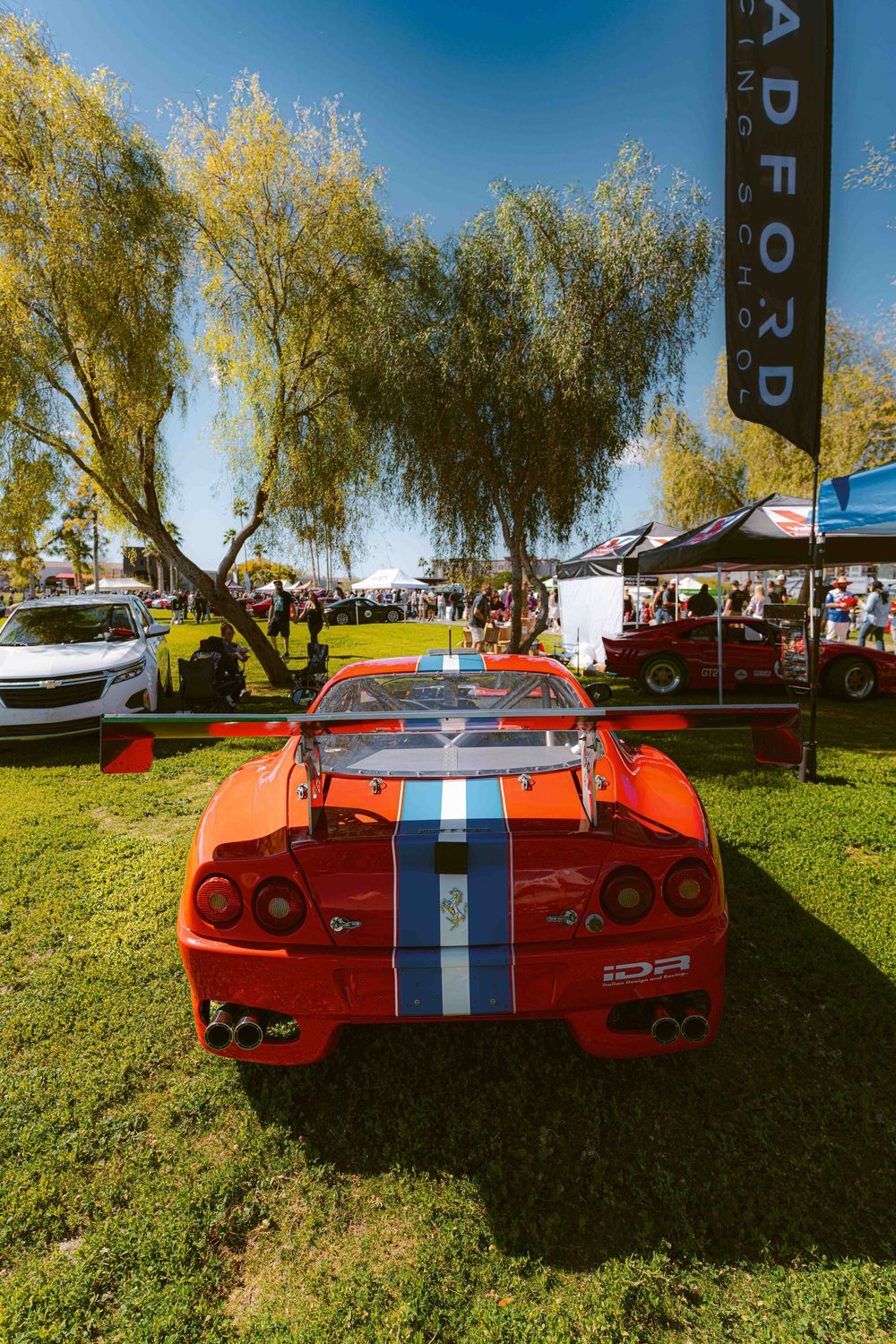 Red Ferrari with white racing stripes displayed at an outdoor car show under willow trees on a sunny day.