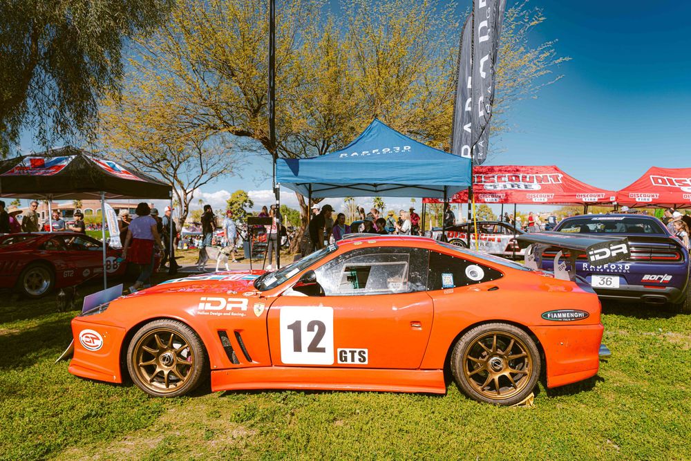 Orange sports cars lined up at an outdoor automotive event under blue skies and trees.