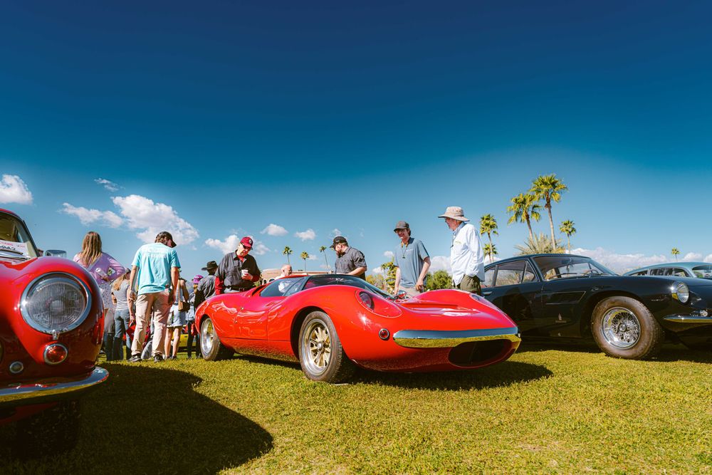 Panoramic view of vintage sports cars lined up at an outdoor car show against bright blue sky.