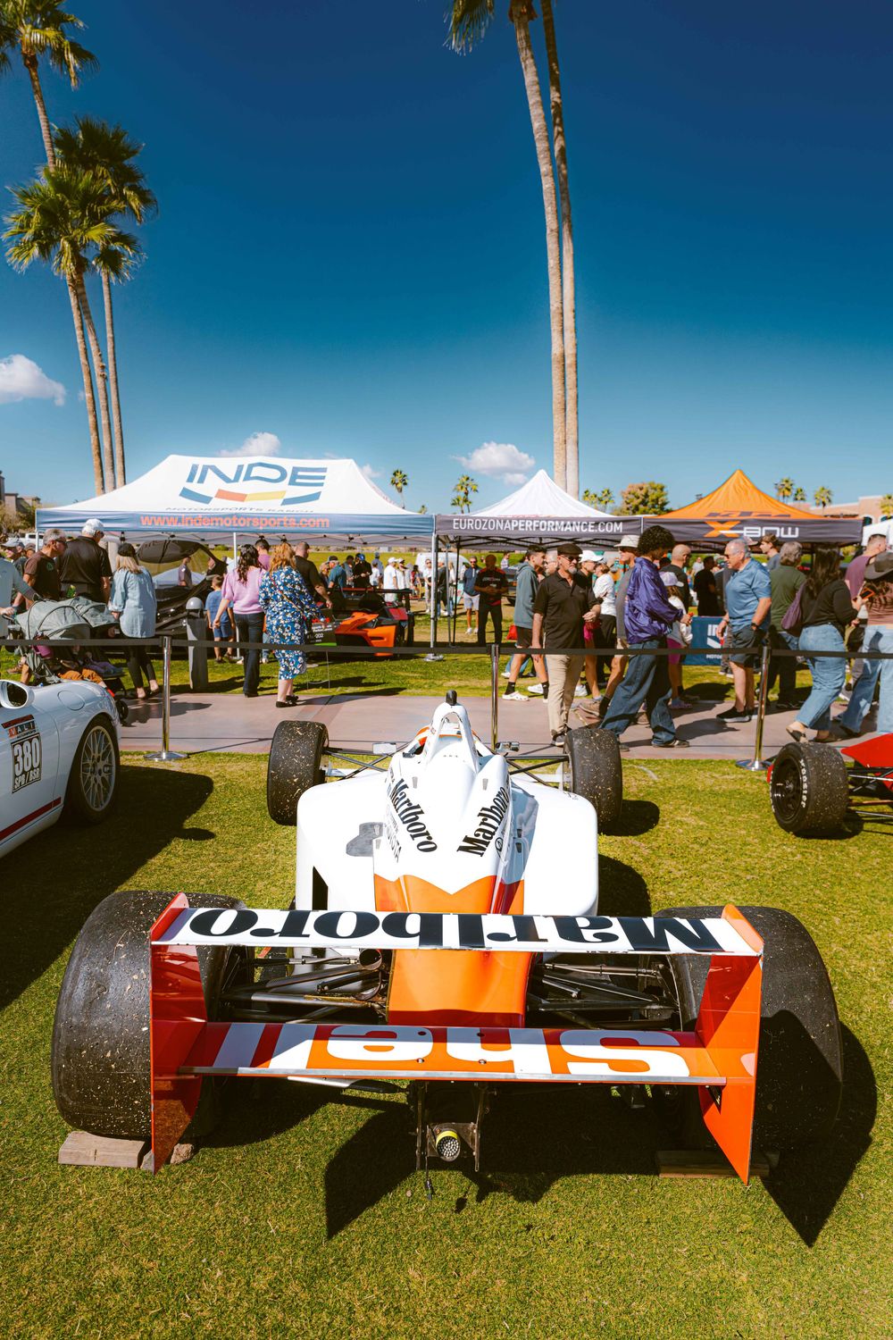 IndyCar race cars on display at an outdoor event with palm trees and blue sky in the background.