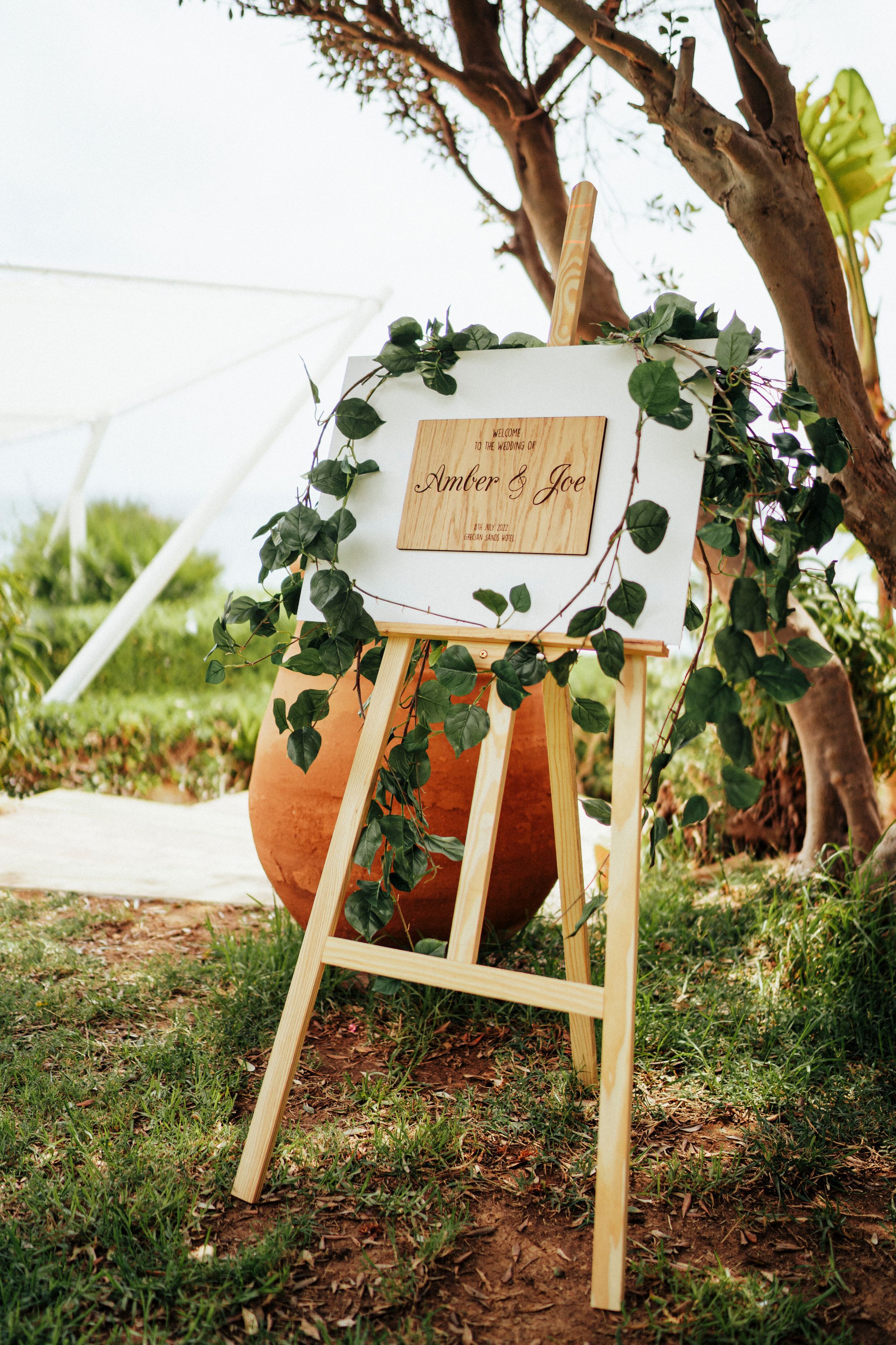 Rustic wooden signs with greenery decorations displayed on wooden stakes in a garden setting.