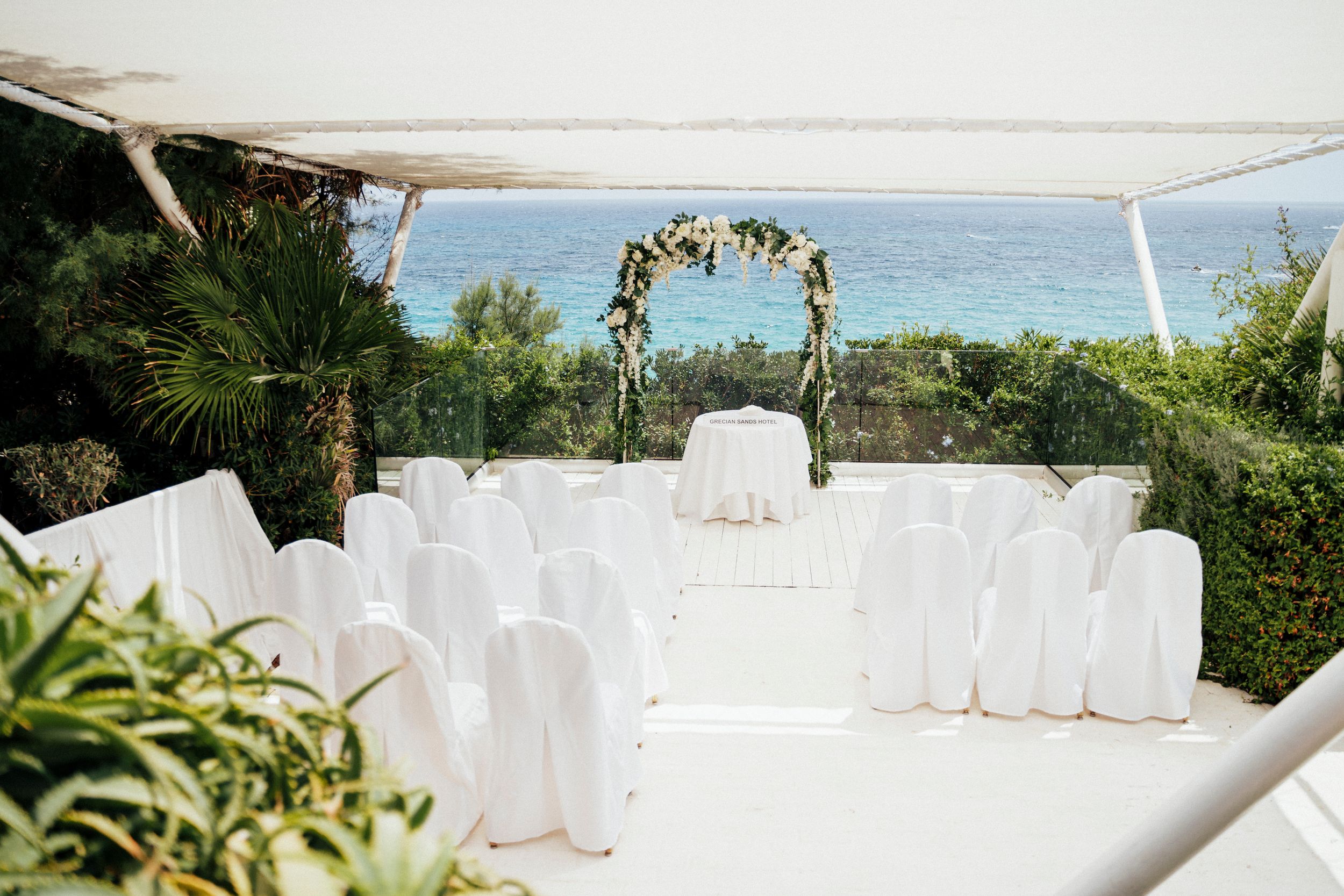 White chairs and tables are set up for an elegant outdoor wedding ceremony with ocean views in the background.