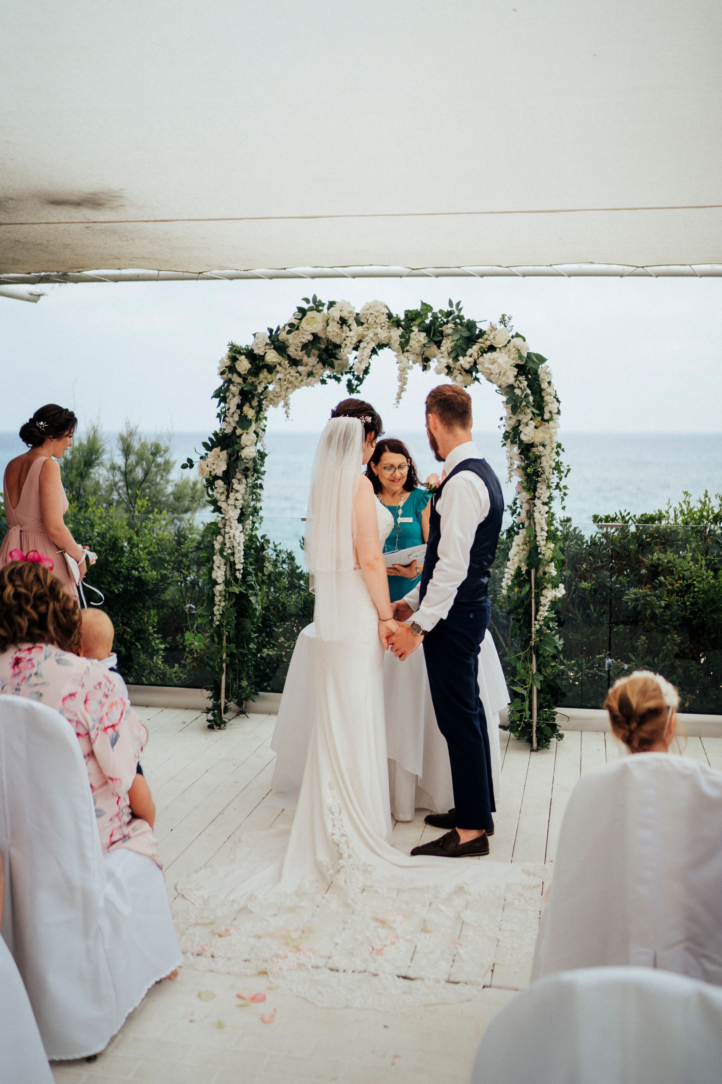 A beachfront wedding ceremony setup with a floral arch and white chairs on sandy shores.