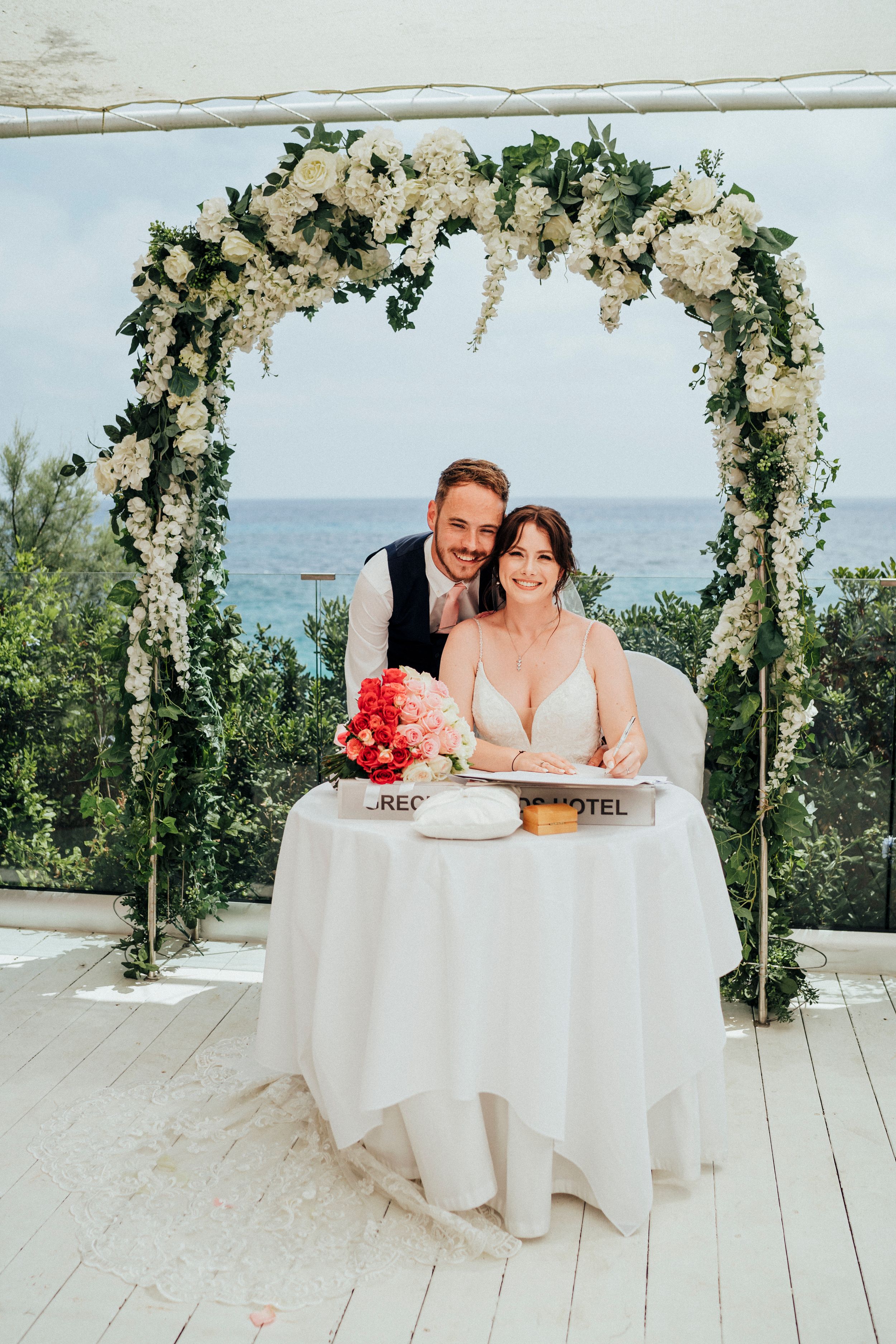 A wedding couple sits at a table under a floral arch with ocean views on an outdoor deck.
