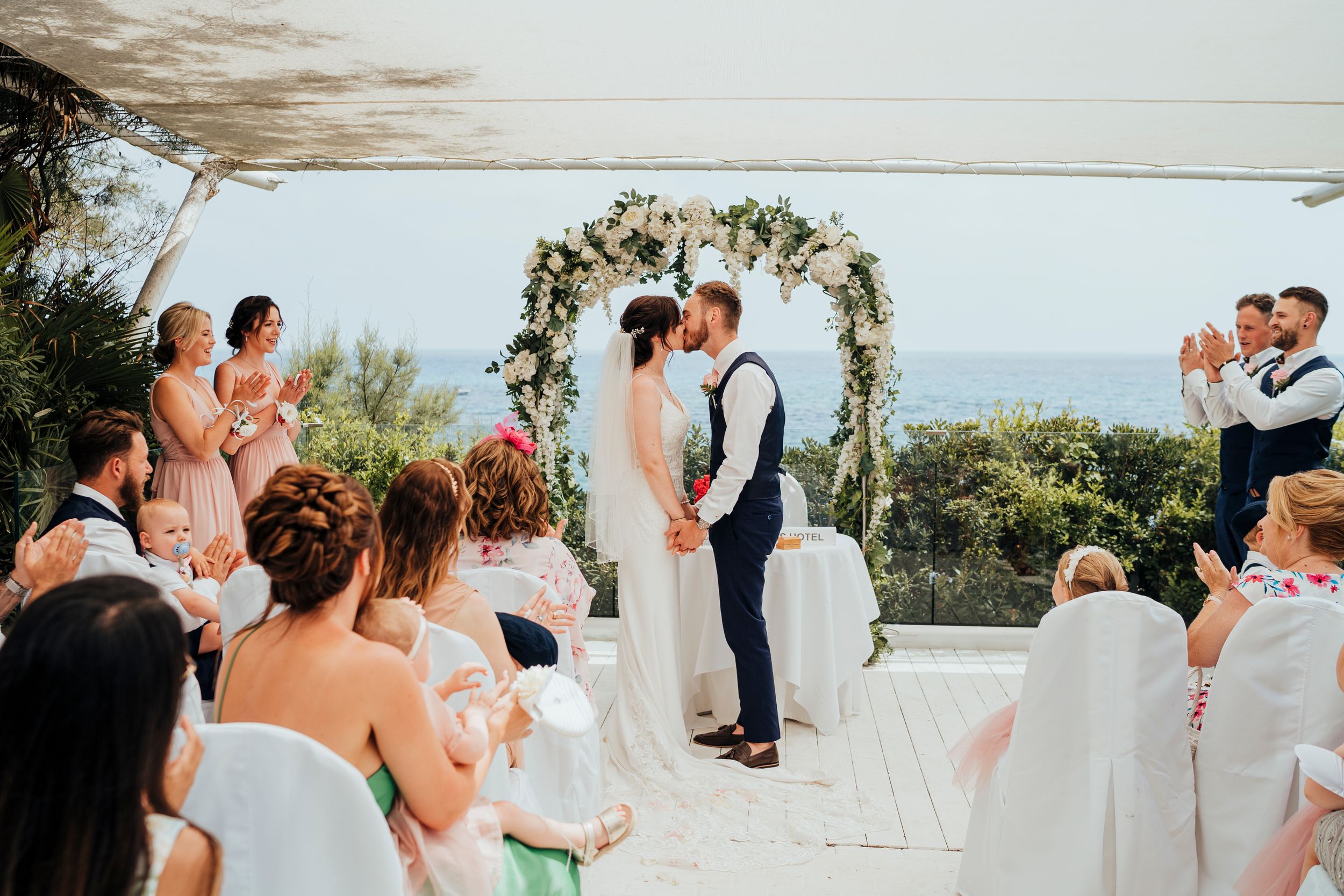 A romantic outdoor wedding ceremony takes place under a floral arch with guests seated on white chairs by the ocean.