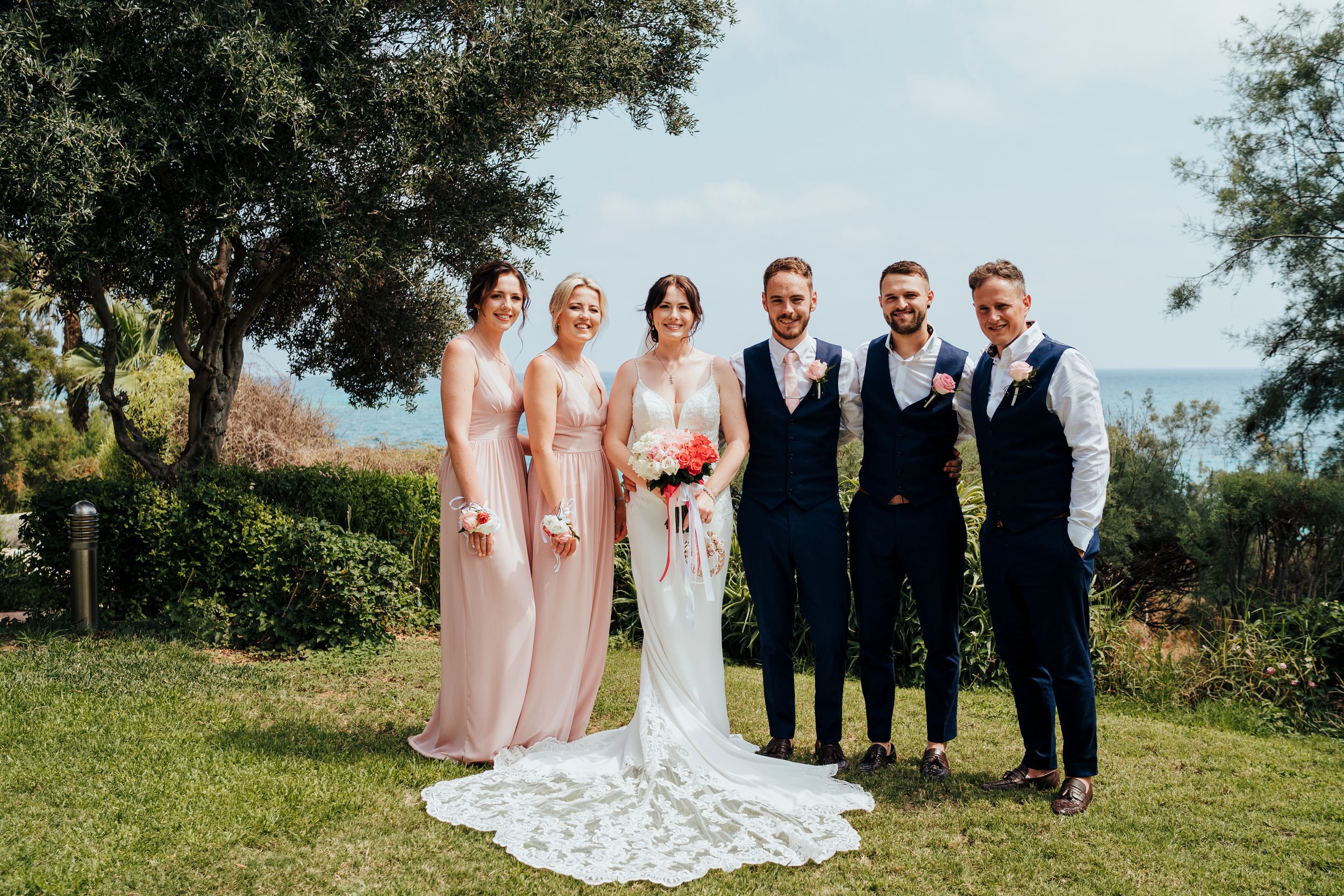 A wedding party poses outdoors in formal attire with bridesmaids in pink dresses and groomsmen in dark suits.