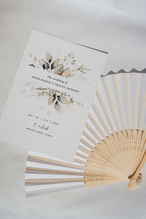 Elegant white paper hand fan with delicate floral design and wedding invitation details.