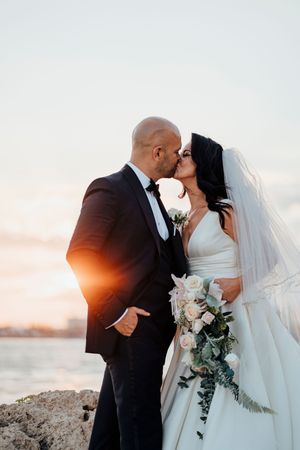 Romantic sunset silhouettes on a rocky beach with flowing white dress and bouquet.