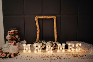 Wedding decoration featuring illuminated 'MR & MRS' letters, an ornate gold frame, and cake display against dark wall.