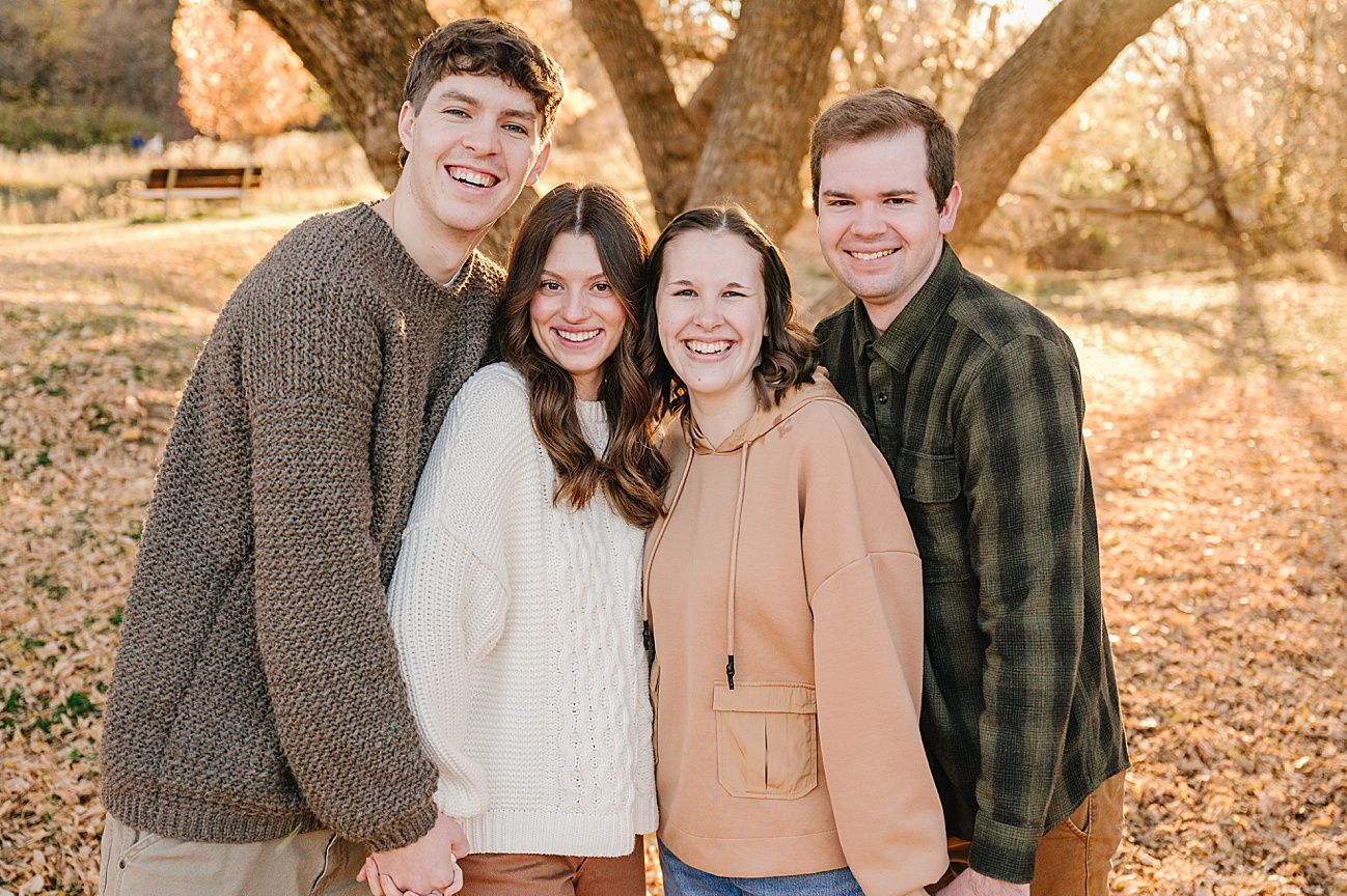 A family of four poses together outdoors on a fall day with autumn leaves on the ground and trees in the background.
