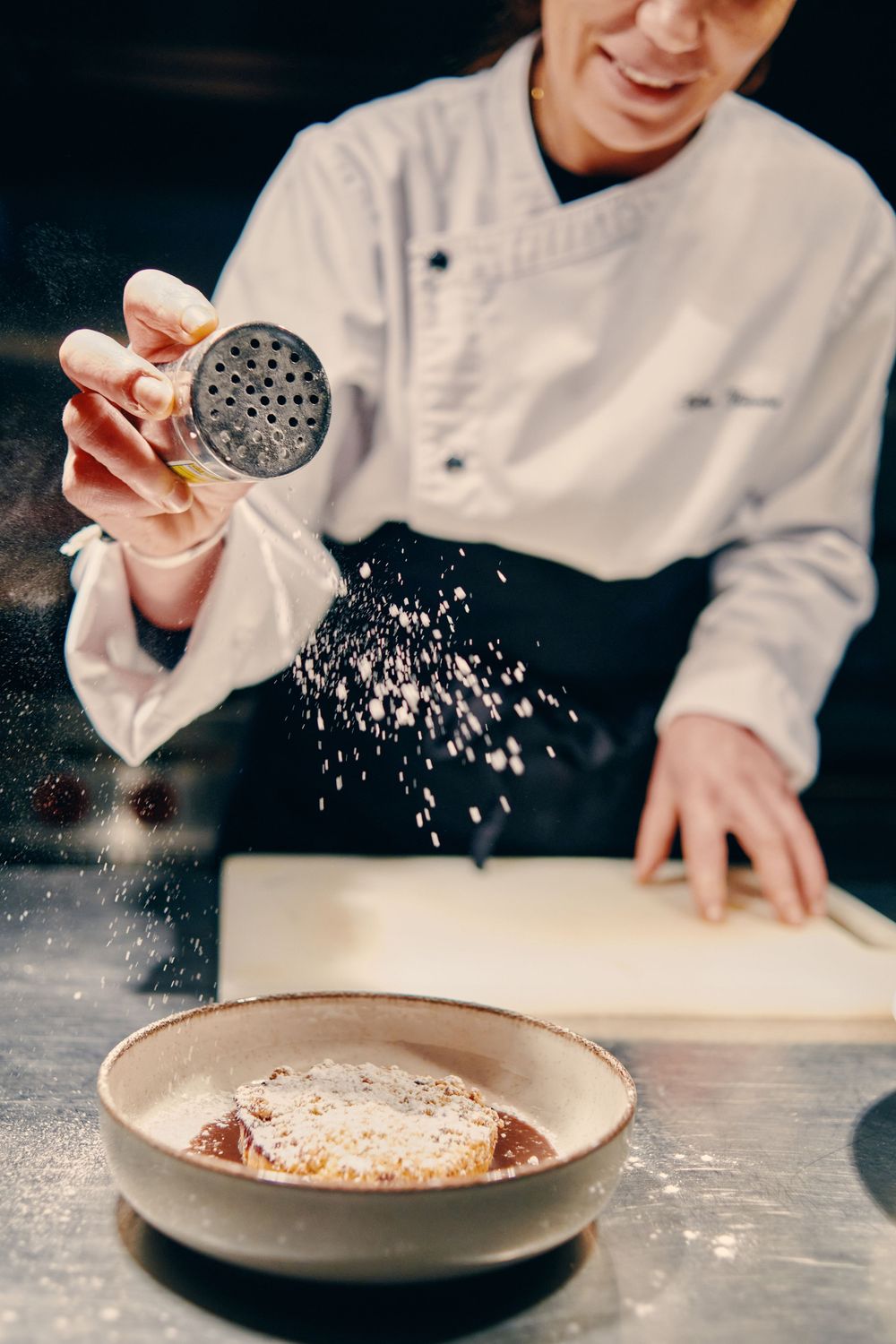 A chef dusting powdered sugar over a dessert in a ceramic bowl in a restaurant kitchen.