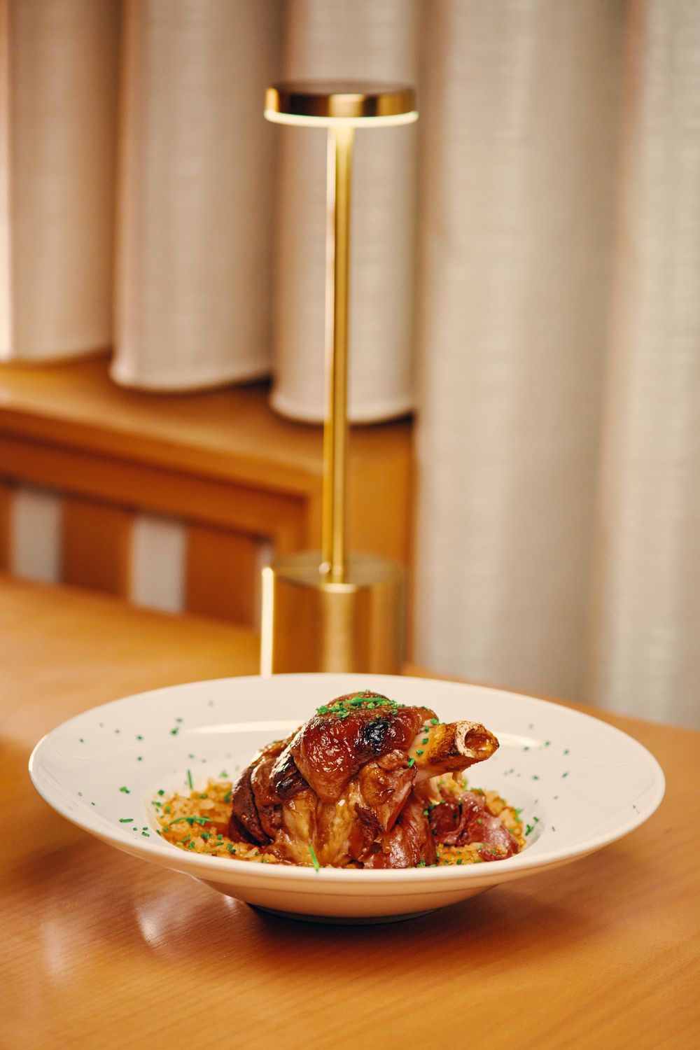 Glazed braised meat dish served on white plate with golden lamp accent in a warm restaurant setting.