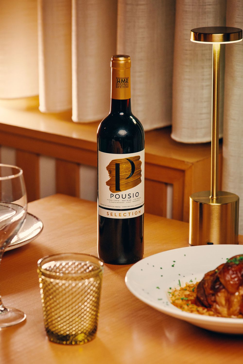 Bottle of Pousio Selection red wine on wooden restaurant table beside a gold lamp and plated dish.