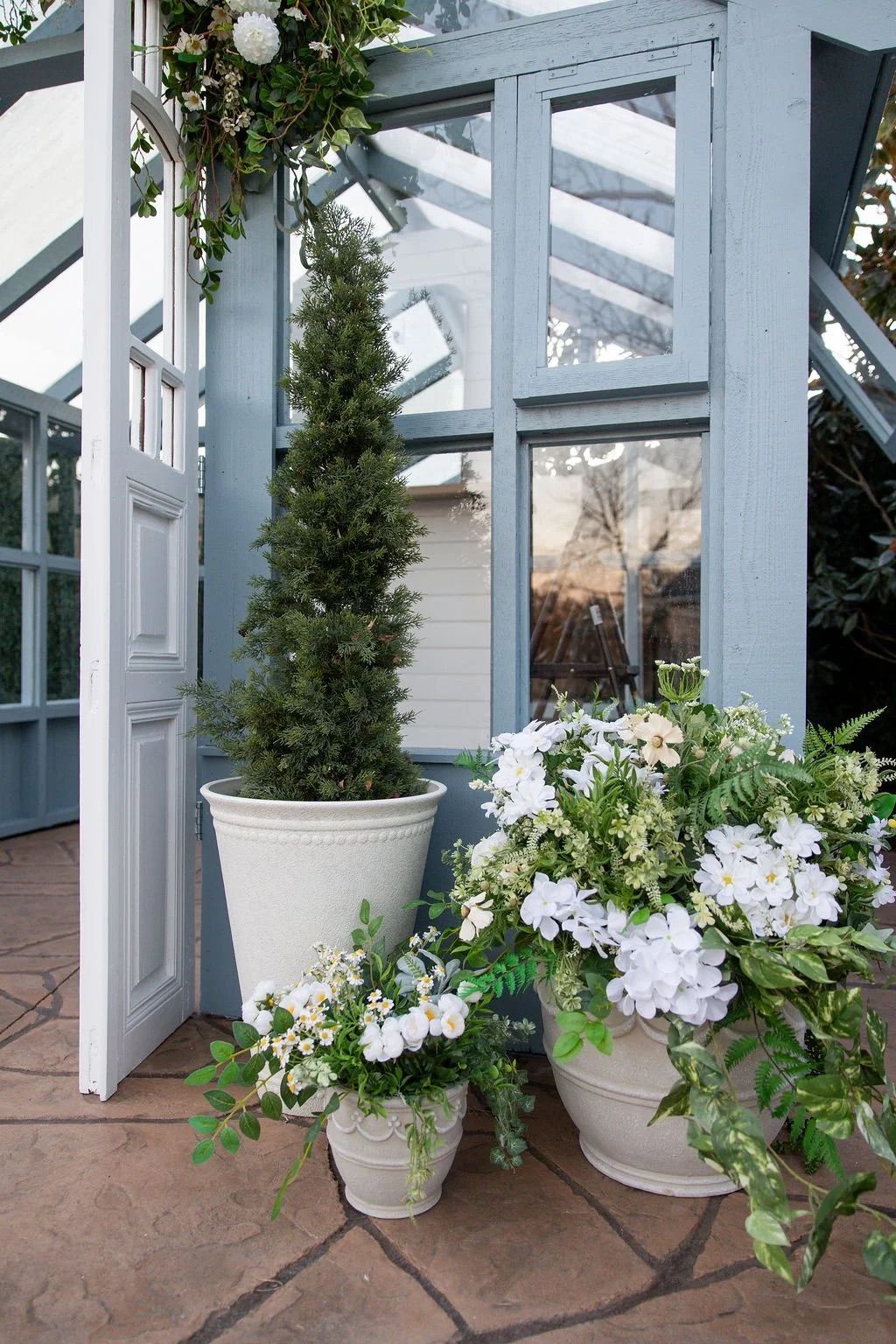 White potted flowers and evergreen plants line a covered patio with white architectural beams and stone flooring.