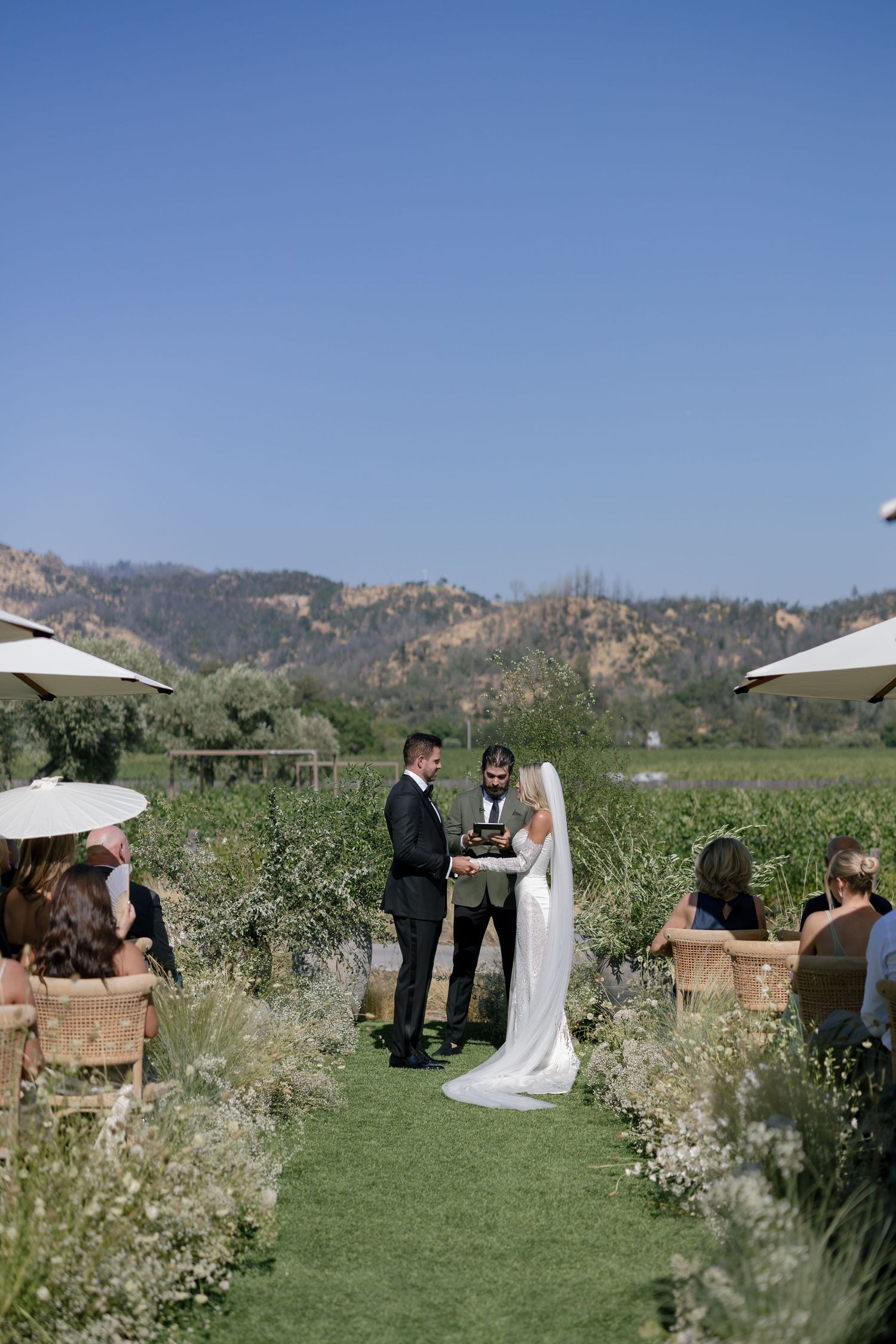 An outdoor wedding ceremony takes place in a scenic mountain setting with green grass and wildflowers.