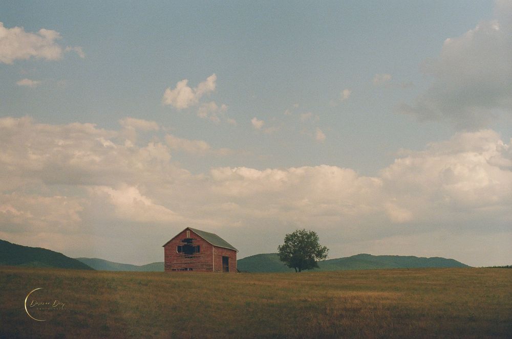 A rustic red barn sits alone on a rolling green hillside beneath a dramatic cloudy sky at sunset in Elkton Virginia